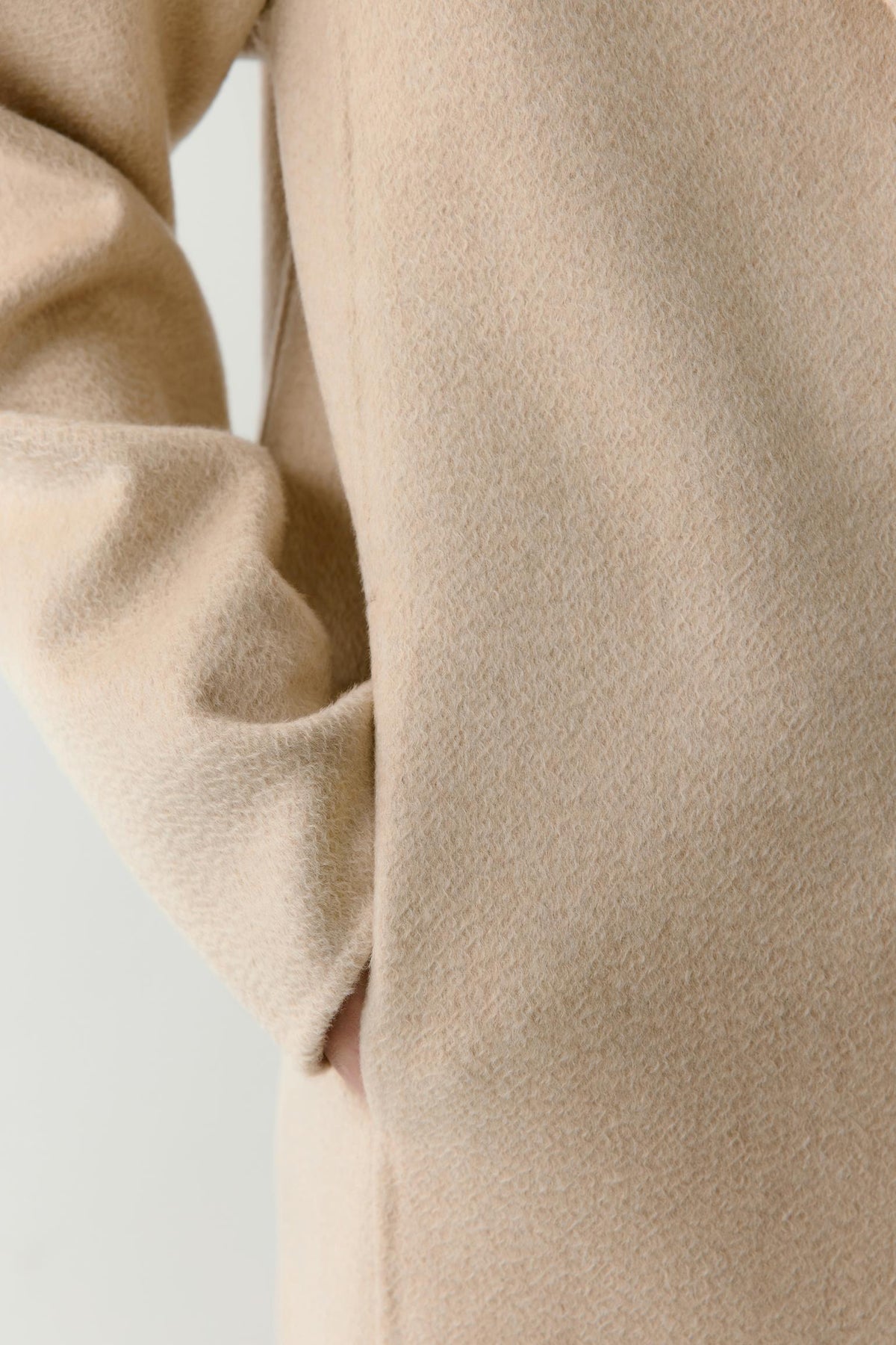 Close-up of a beige wool or cashmere coat sleeve with a soft, textured surface and a hand partially tucked into a side pocket.
