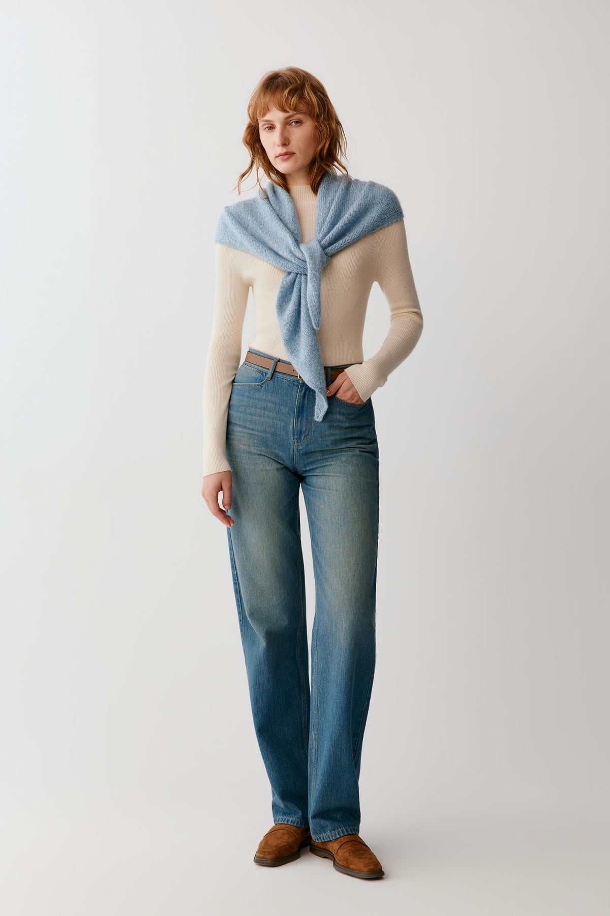 A model stands against a light gray studio background wearing a cream ribbed knit sweater, blue jeans, brown suede shoes, and a light blue knit wrap tied around her shoulders.