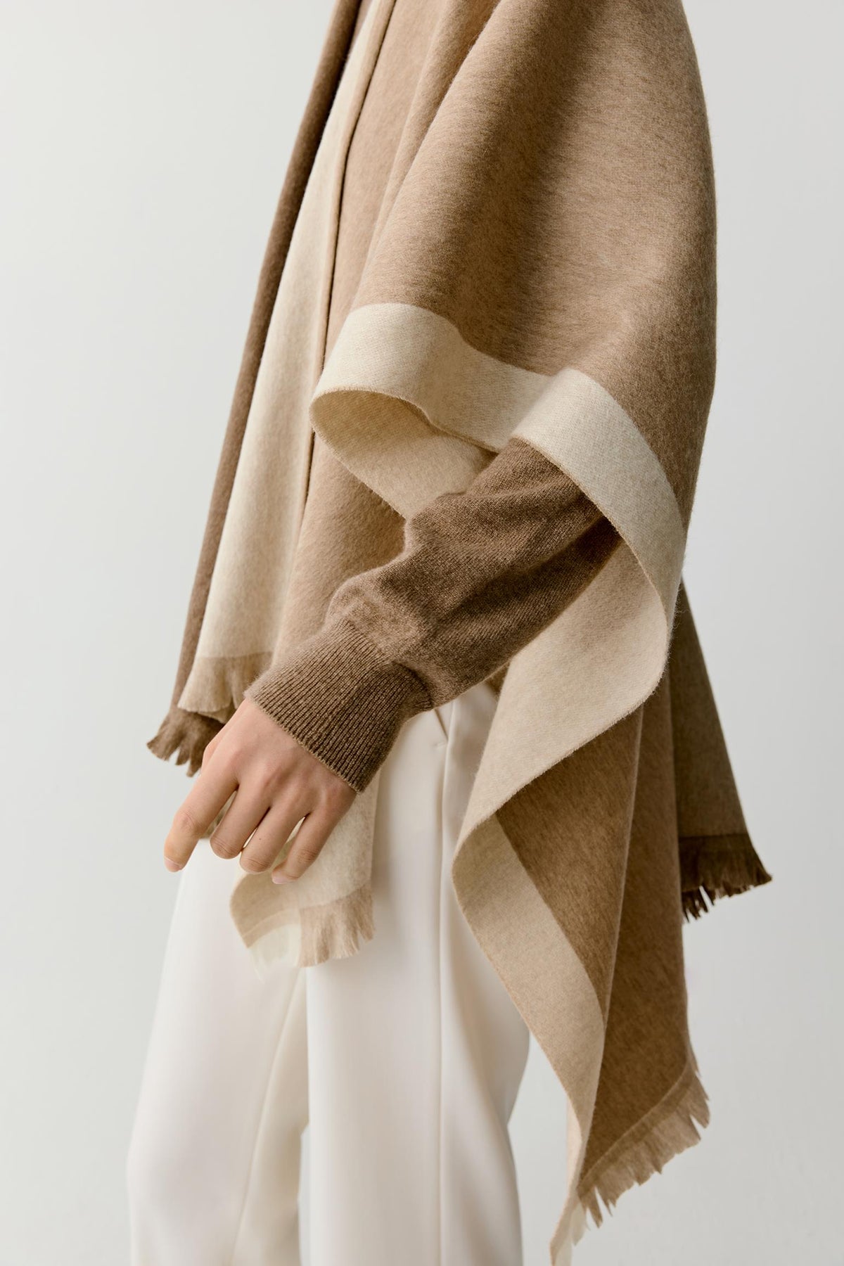A close-up of a person wearing a draped camel brown and beige shawl over a brown knit sweater and white trousers, against a plain light gray background.