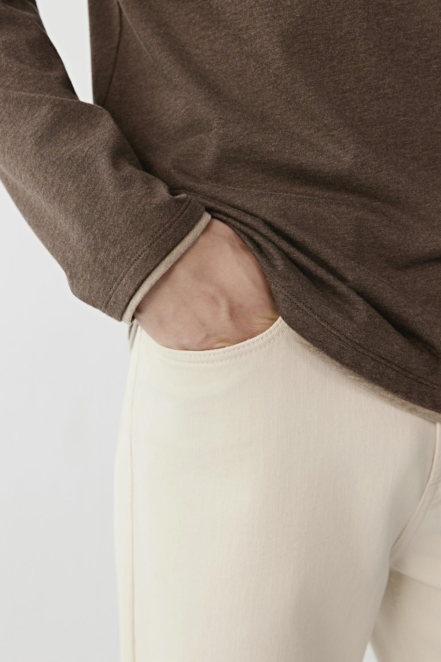 Close-up of a person wearing a brown long-sleeve top and off-white pants, with the hand tucked into the pocket of the pants.