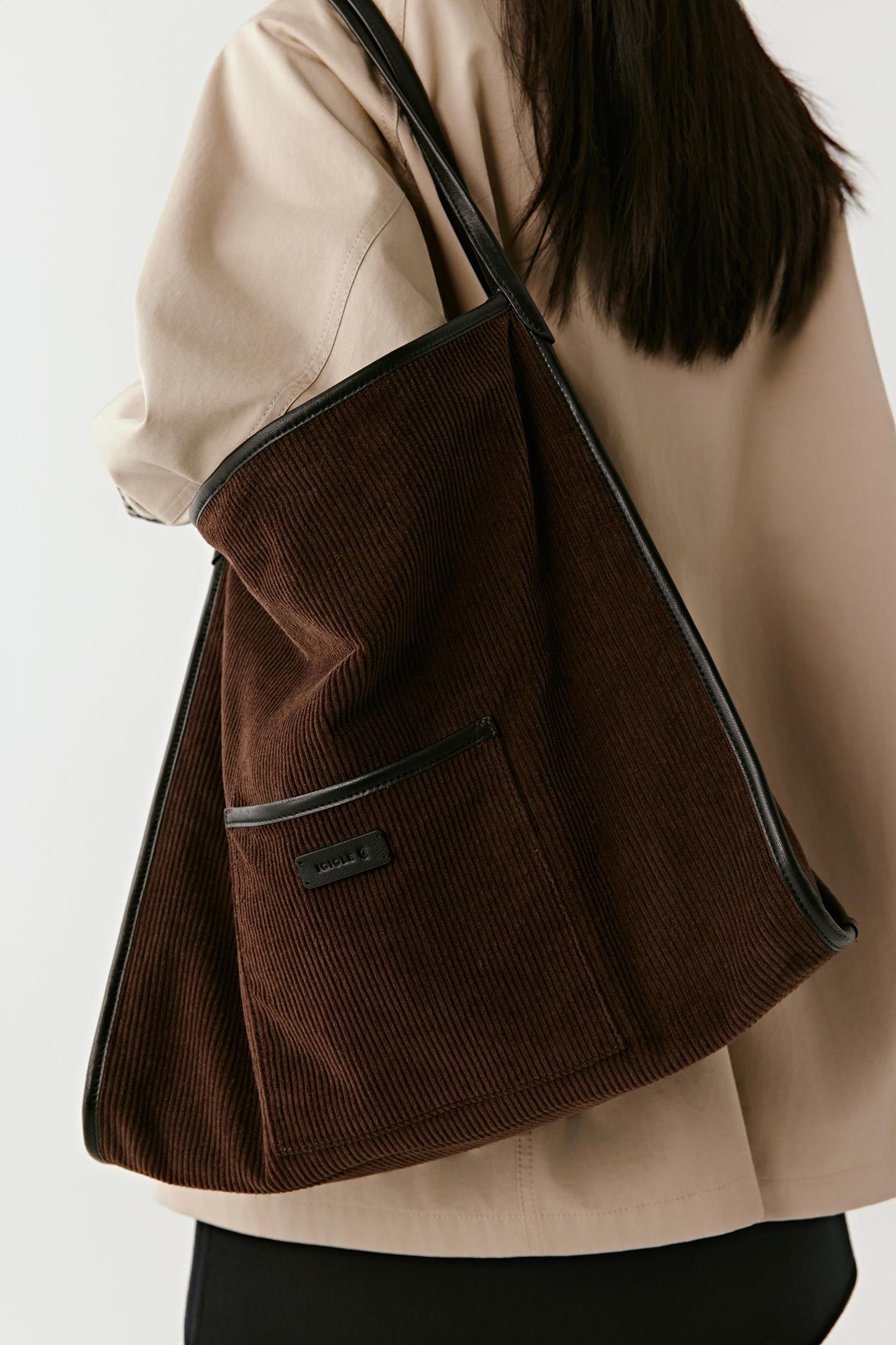 A dark brown corduroy tote bag with black trim and a shoulder strap, worn over the shoulder of a person wearing a beige blazer.