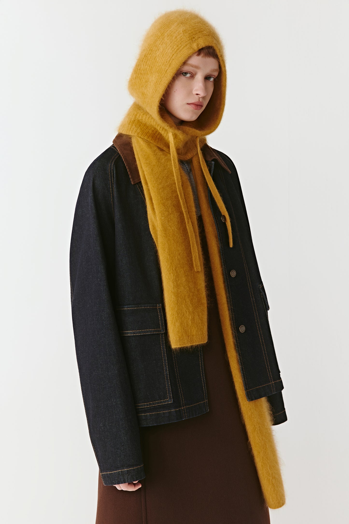 Woman wearing a mustard yellow knit hood with long drawstrings styled with a black denim jacket on a light background