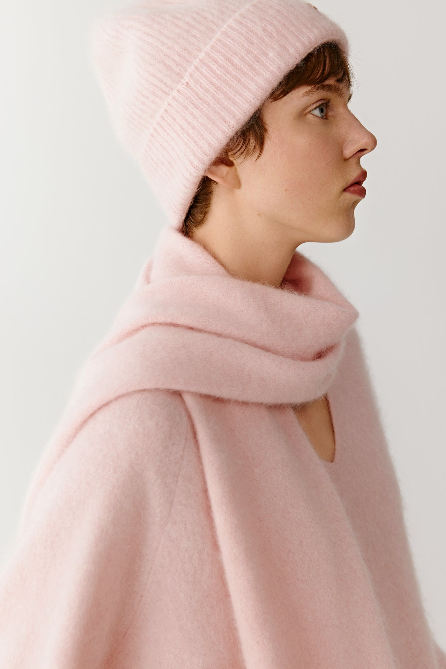 Side view of a woman wearing a pink knit beanie and scarf with a casual pink sweater on a light background