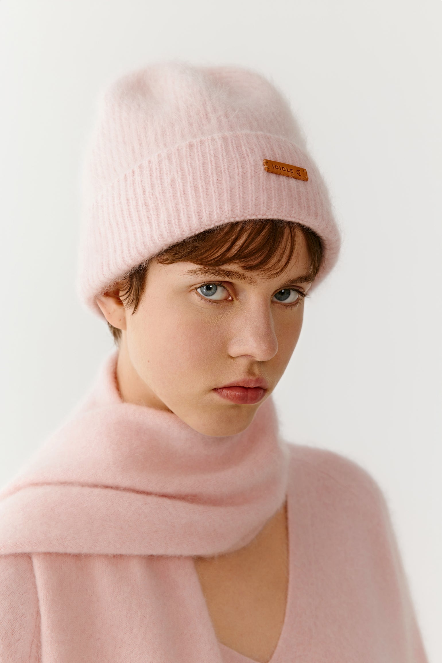 Woman wearing a light pink knit beanie with a subtle logo and matching scarf on a light background