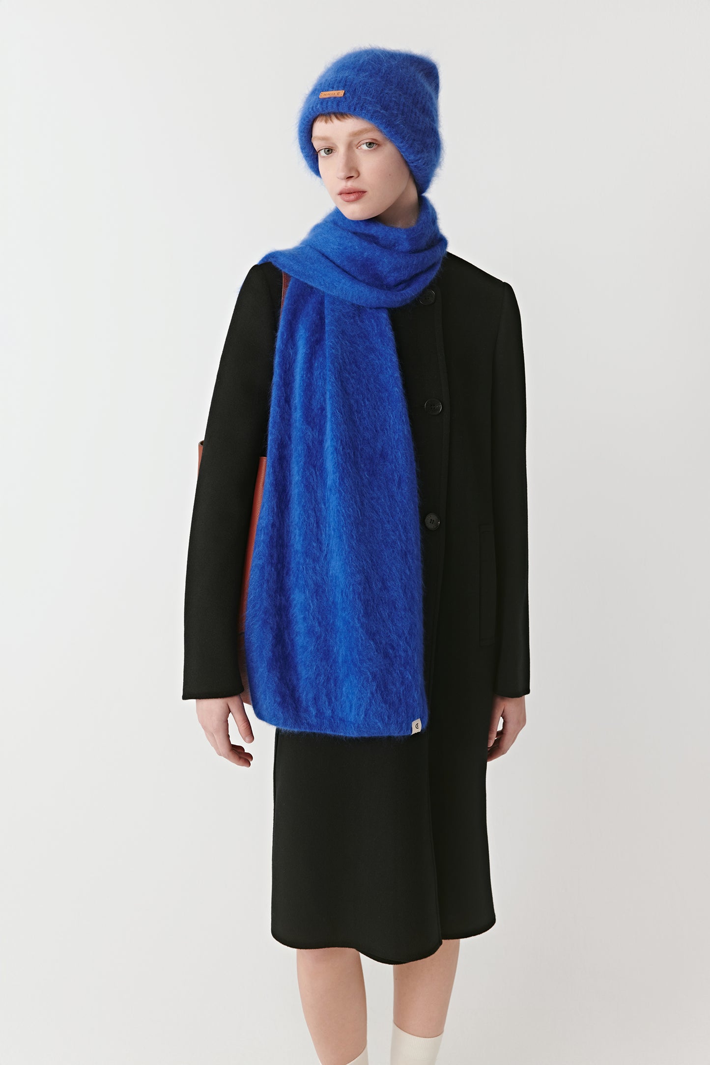 Full body view of a woman wearing a bright blue knit beanie and scarf on a light background