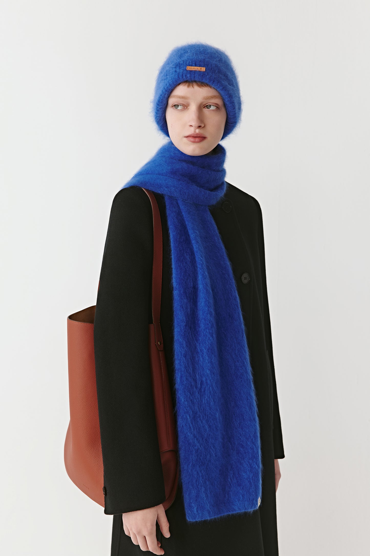 Woman wearing a vibrant blue knit beanie and matching scarf styled with a black coat on a light background