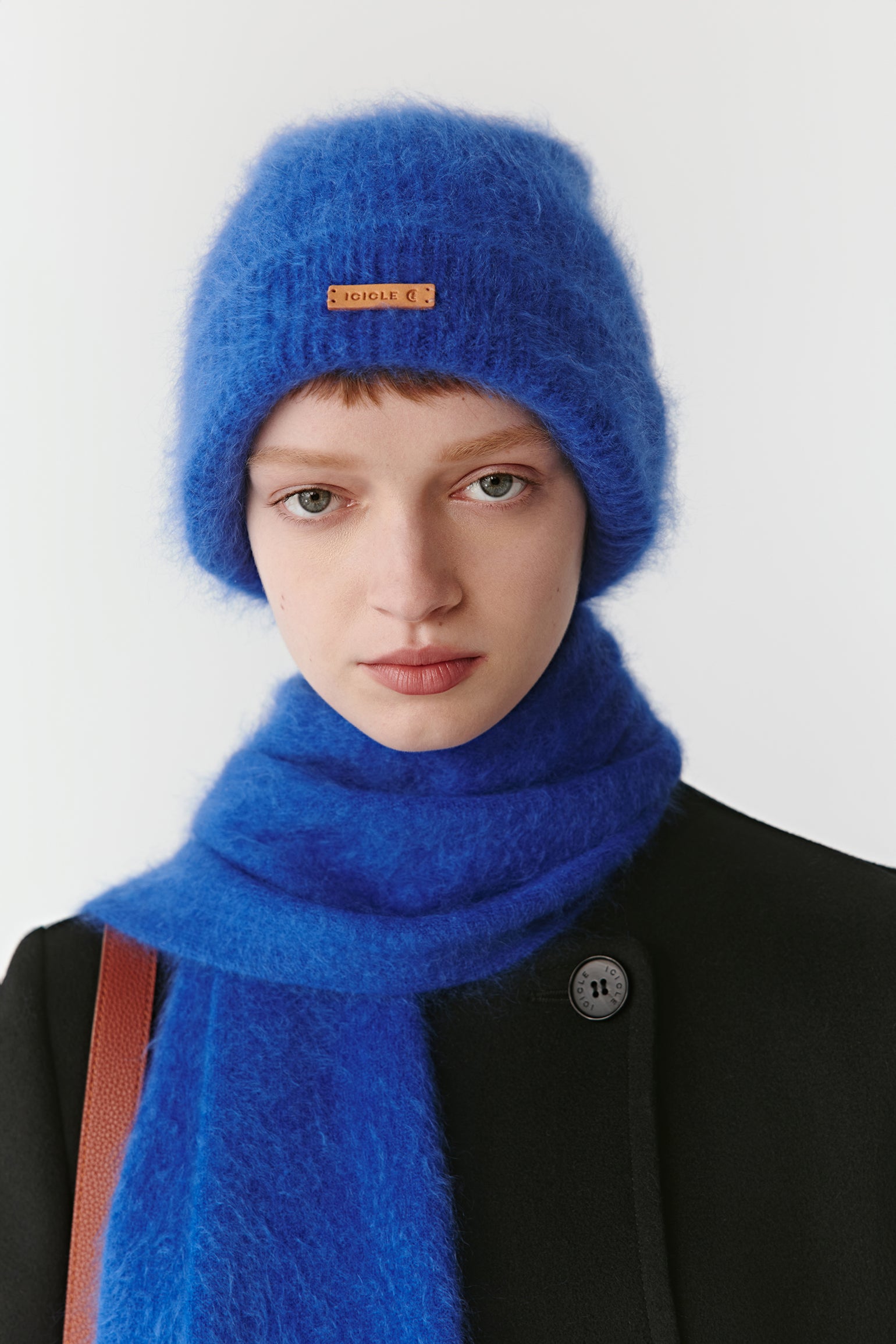 Woman wearing a bright blue knit beanie and matching scarf styled with a black coat on a light background