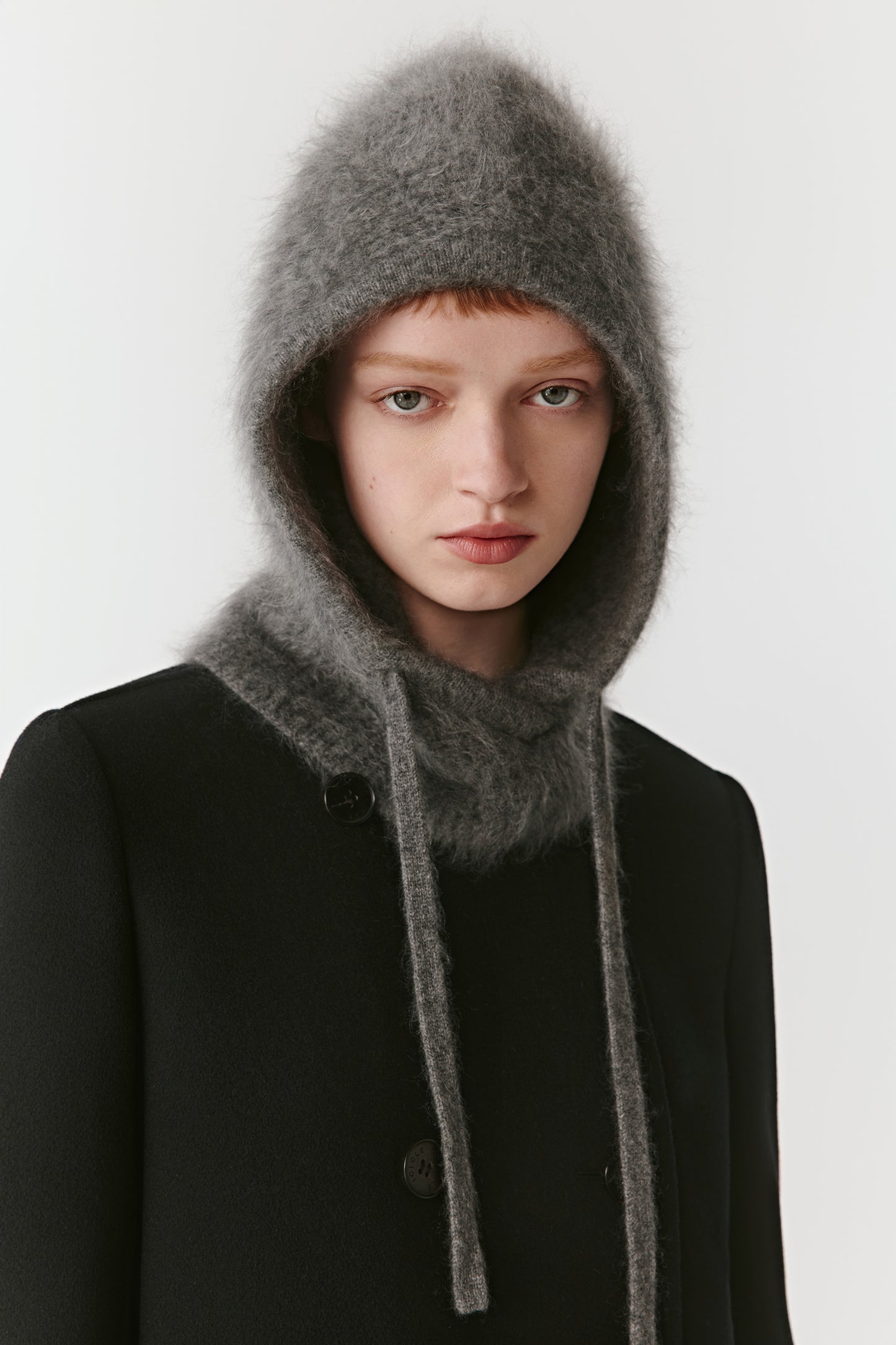 Woman wearing a grey knit hood with long drawstrings styled with a black coat on a light background