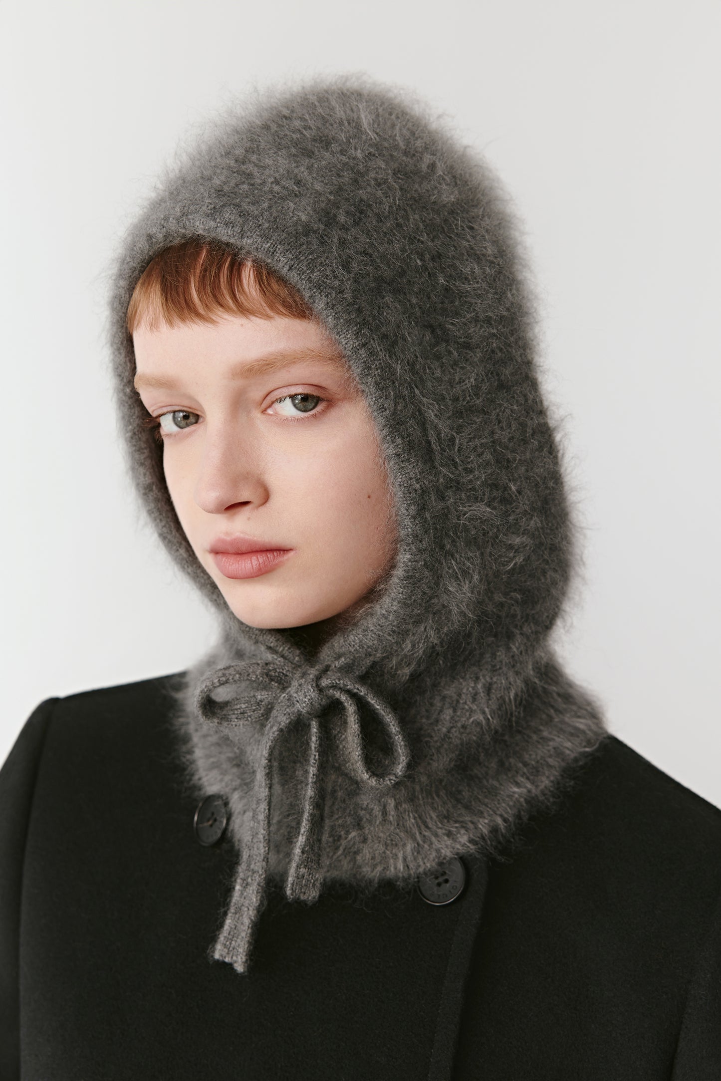 Side view of a woman wearing a grey knit hood with long ties on a light background