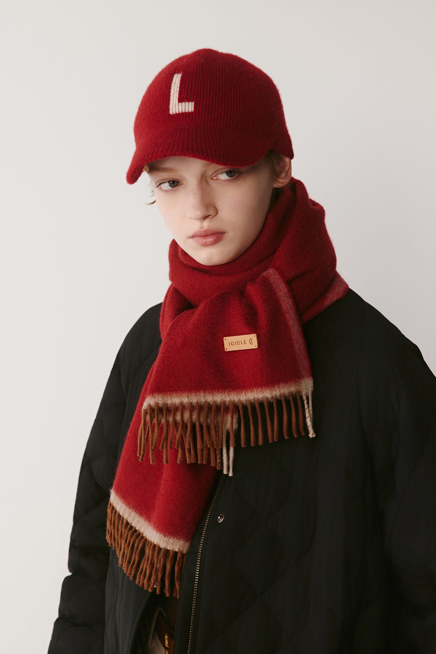 Woman wearing a red knit scarf with fringed edges and a matching burgundy cap over a black coat on a light background