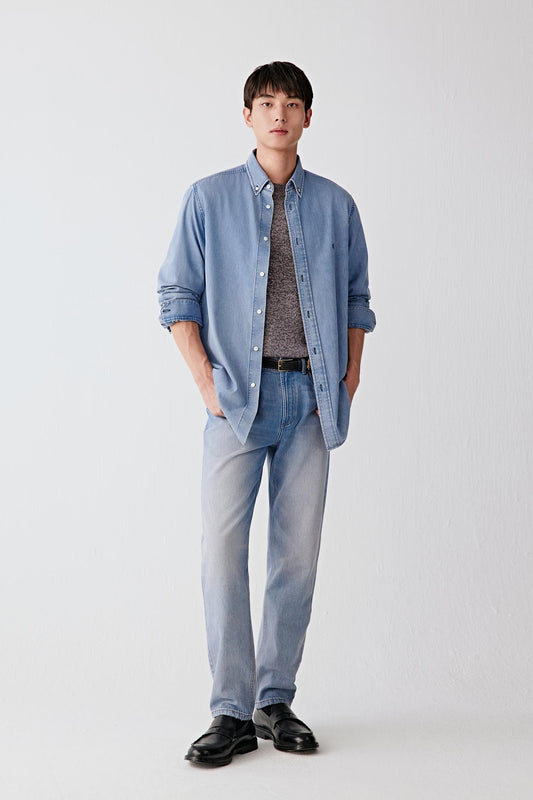 Full-length front view of a men’s light blue cotton denim shirt worn open over a darker T-shirt, paired with light trousers, showing a relaxed straight fit and soft washed texture.