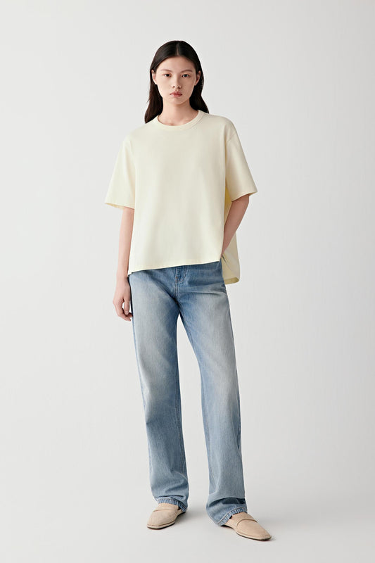 Full-length view showing the T-shirt paired with light blue denim trousers. The image emphasizes the fluid drape of the cotton jersey, the relaxed fit and the lightweight structure of the garment.