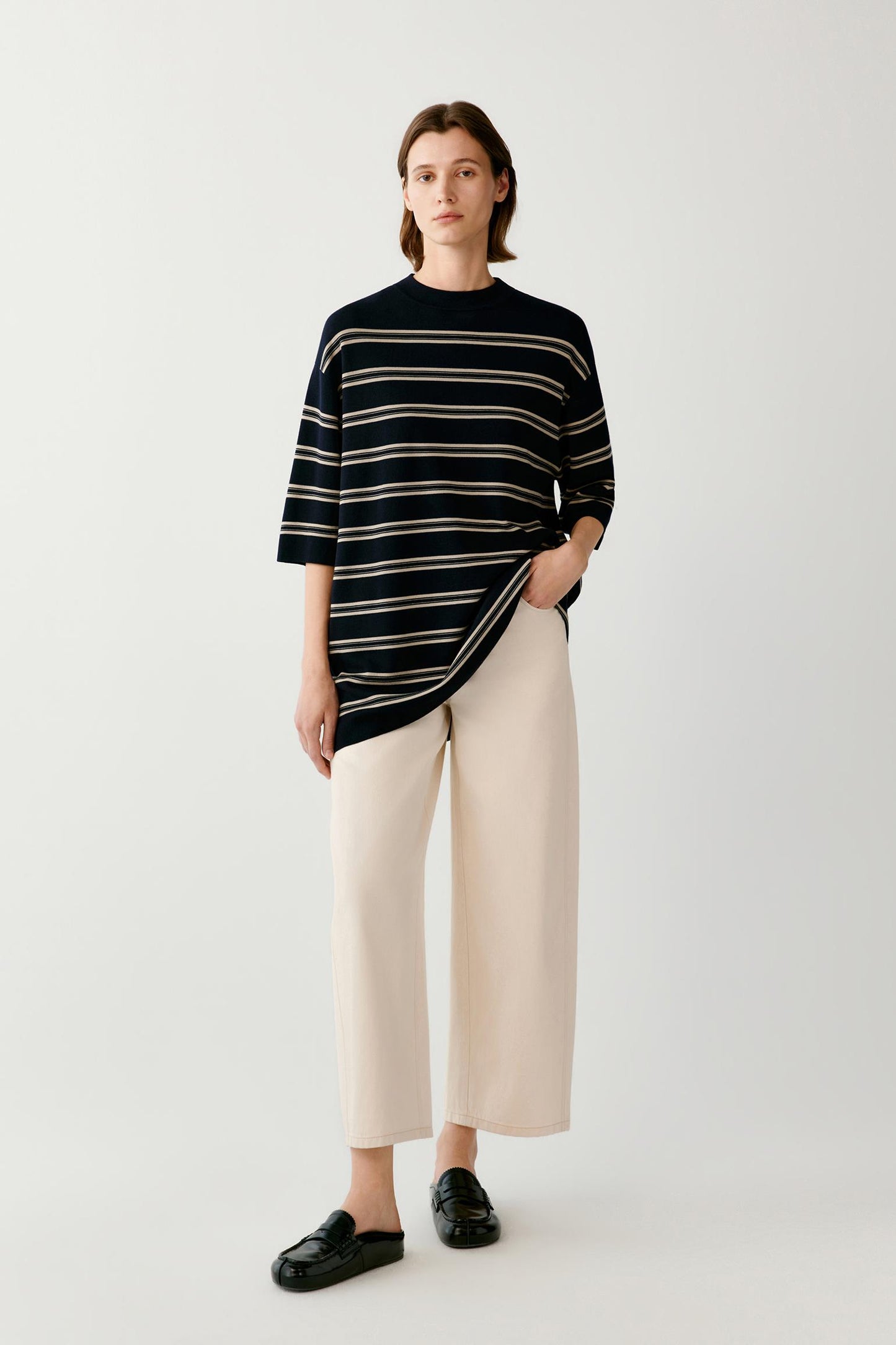 A model stands against a light gray studio background wearing a black knit top with thin cream horizontal stripes, paired with cream wide-leg cropped pants and black loafers.