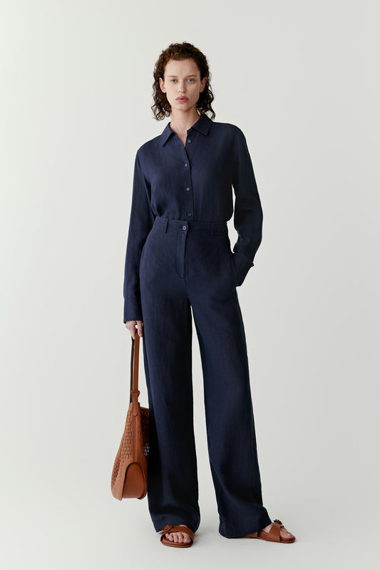 Front view showing the full silhouette and the natural movement of the linen fabric. The image emphasizes the fluid fall of the shirt, the straight cut through the body and the balanced proportions created by the long sleeves and curved hem.