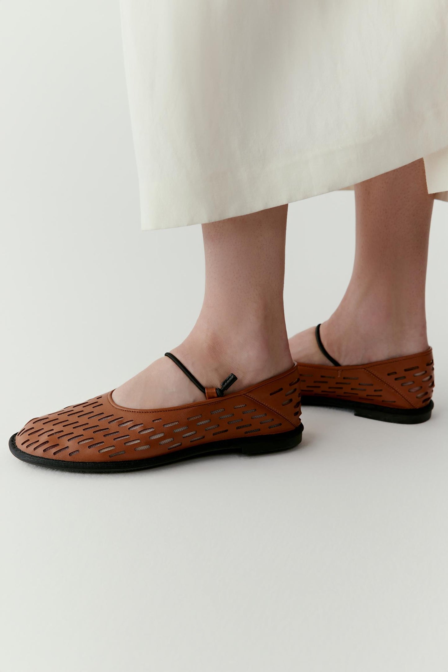 Side view highlighting the flat sole and rounded toe shape. The perforated leather and slender strap emphasize the light, flexible construction of the shoes.