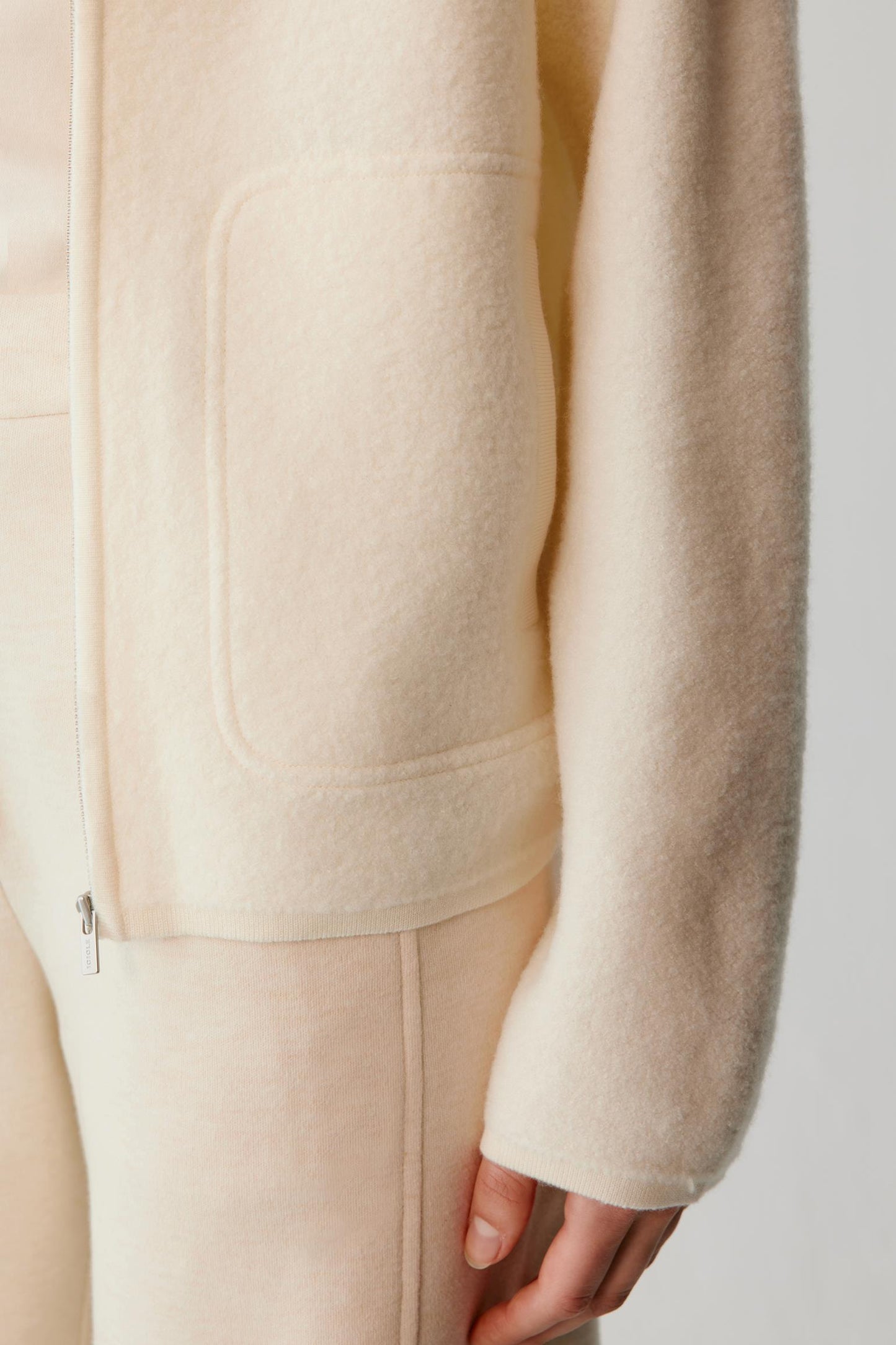 Close-up of a person wearing a soft cream-colored zip-front jacket with a large patch pocket and matching trousers, photographed against a light neutral background.