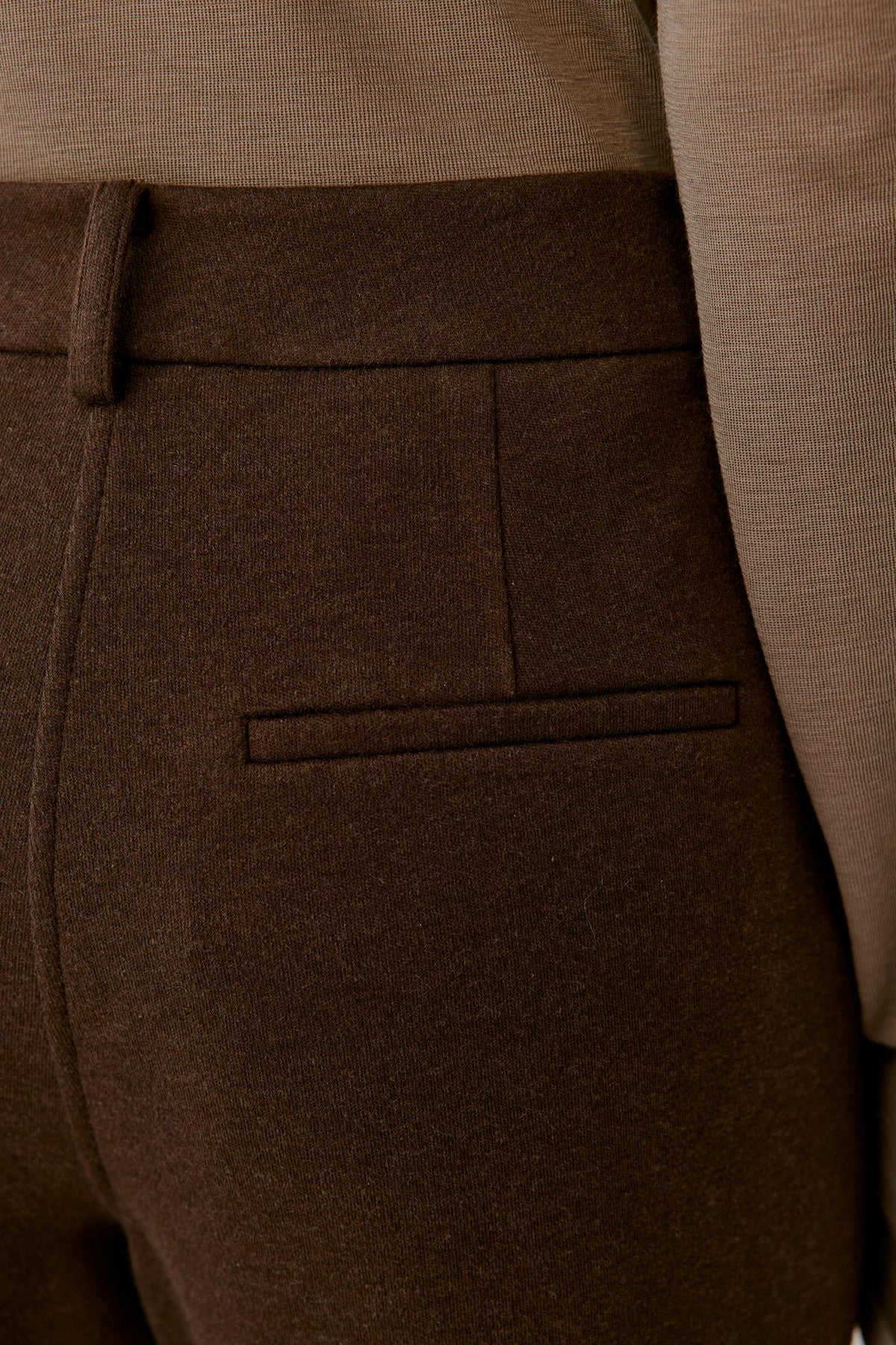 Close-up of dark brown tailored trousers showing the back waistband, belt loop, and a welt pocket, worn with a taupe knit top.
