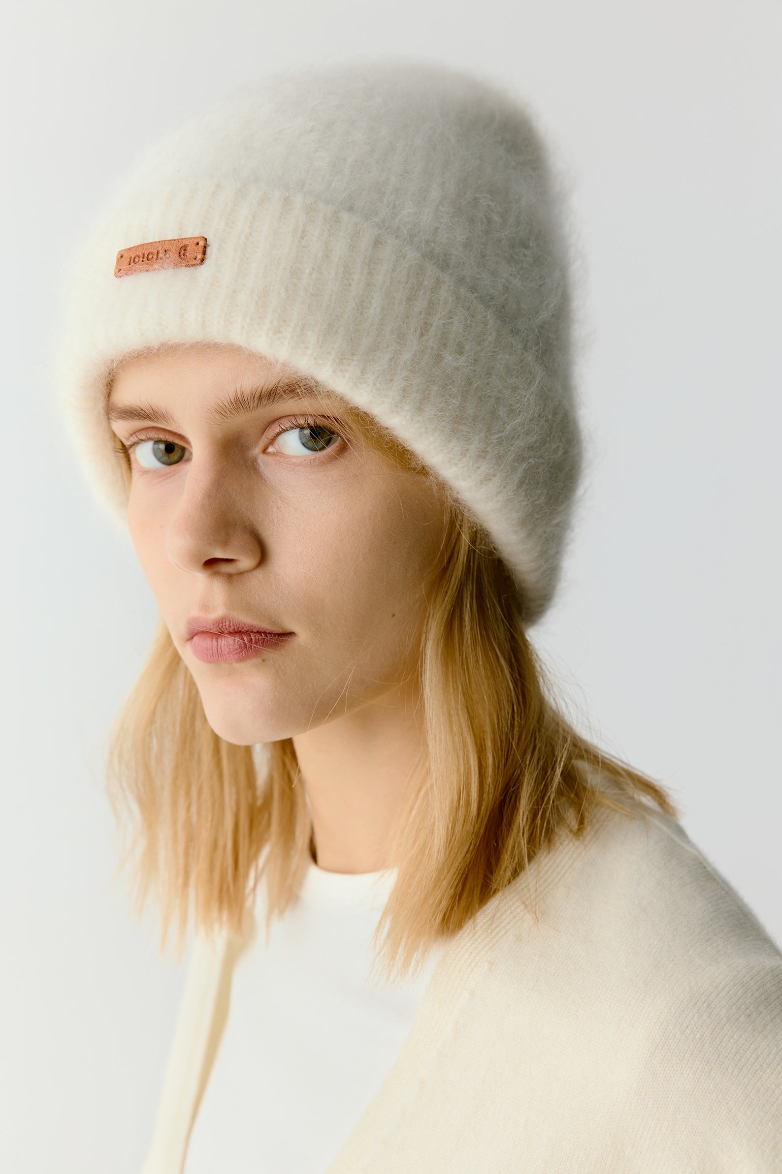 Woman wearing a white knit beanie with a minimal logo on a light background