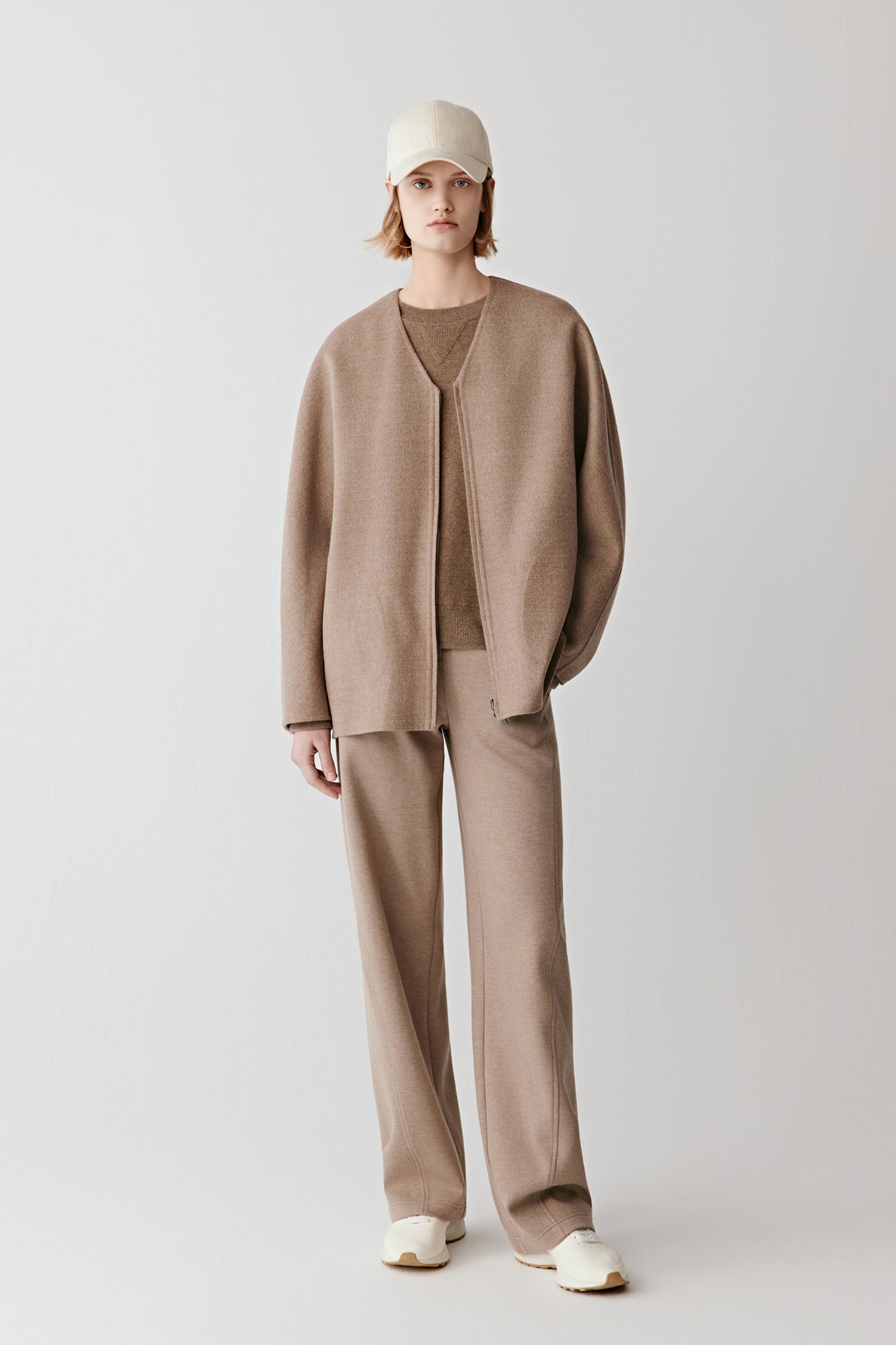 Model standing against a light grey background wearing a short grey-brown double-faced wool and silk coat. Front view showing dropped shoulders, round neckline and a relaxed straight cut paired with tonal trousers.