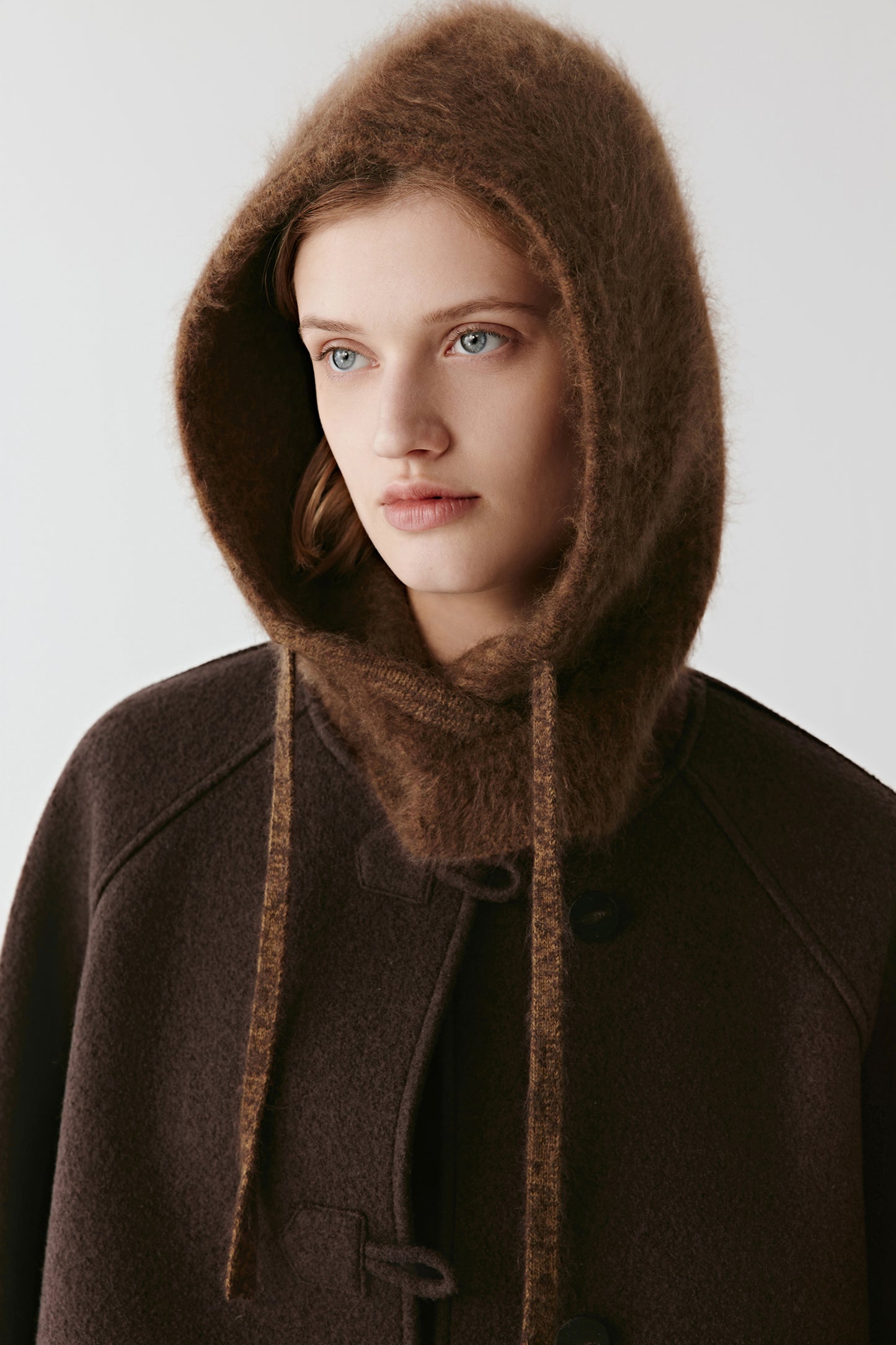 Woman wearing a brown knit hood with long drawstrings styled with a dark coat on a light background