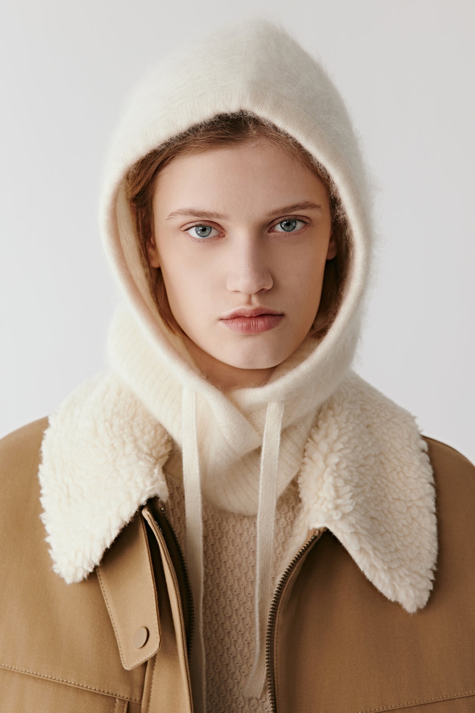 Woman wearing a cream knit hood with long drawstrings styled with a shearling collar coat on a light background