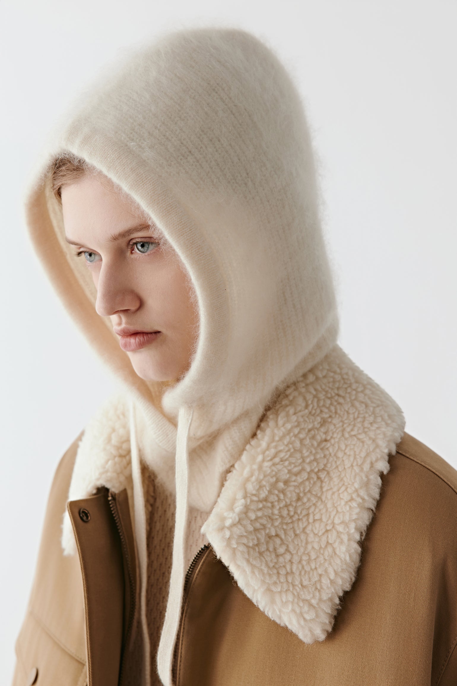 Side view of a woman wearing a cream knit hood with long ties on a light background