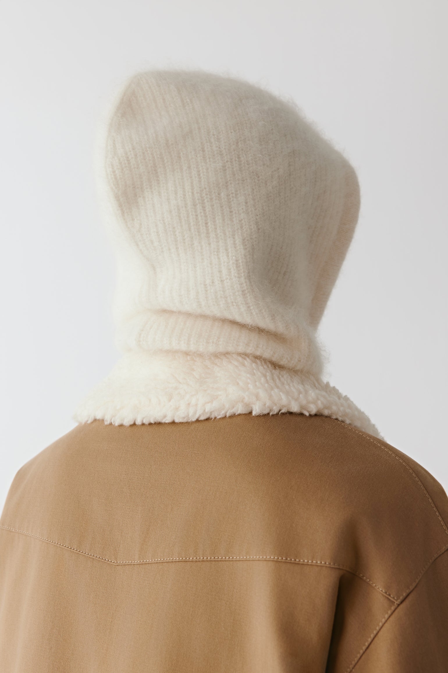 Back view of a cream knit hood with long ties styled with a coat on a light background