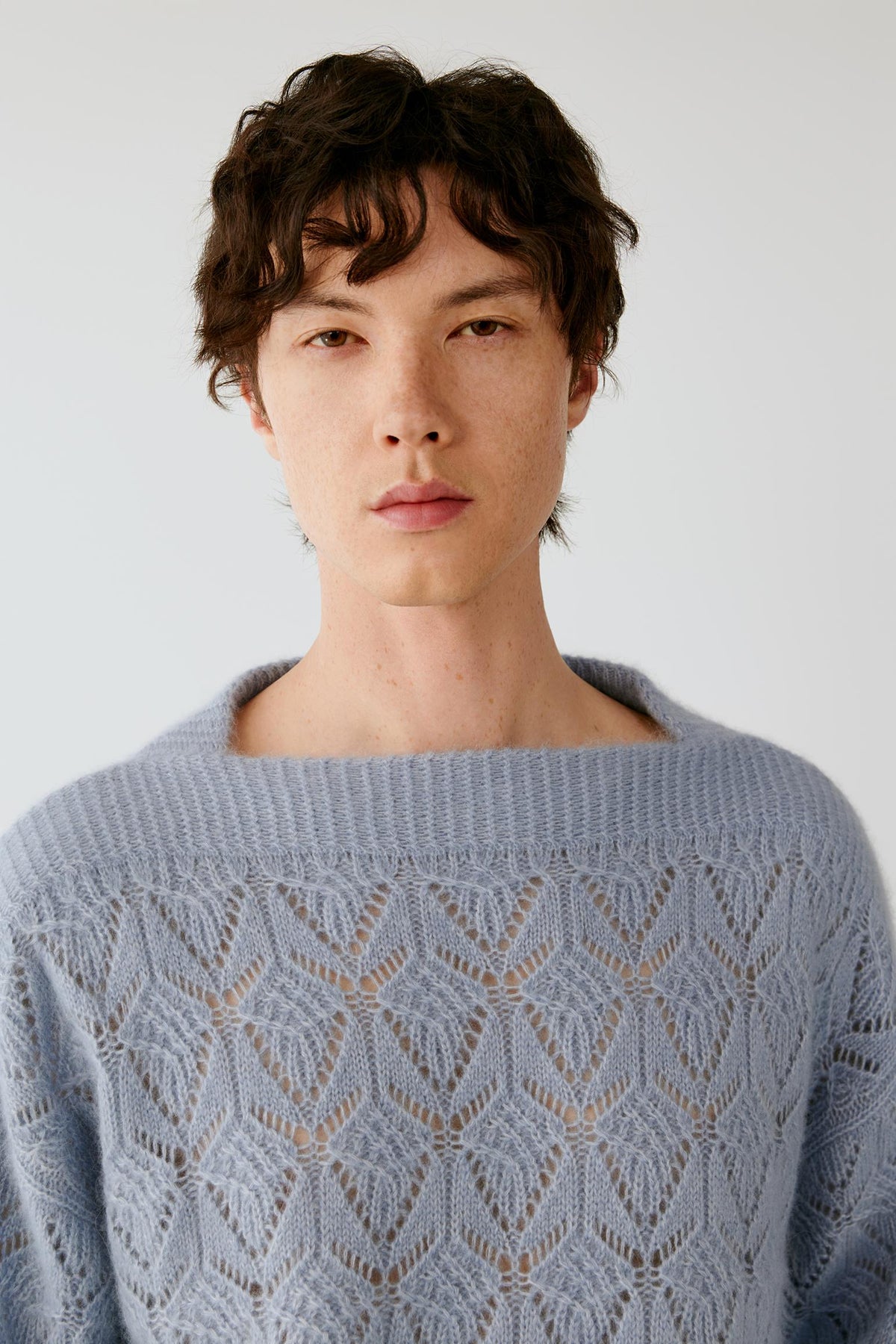 A man wearing a light blue open-knit crewneck sweater, photographed against a plain light gray background.