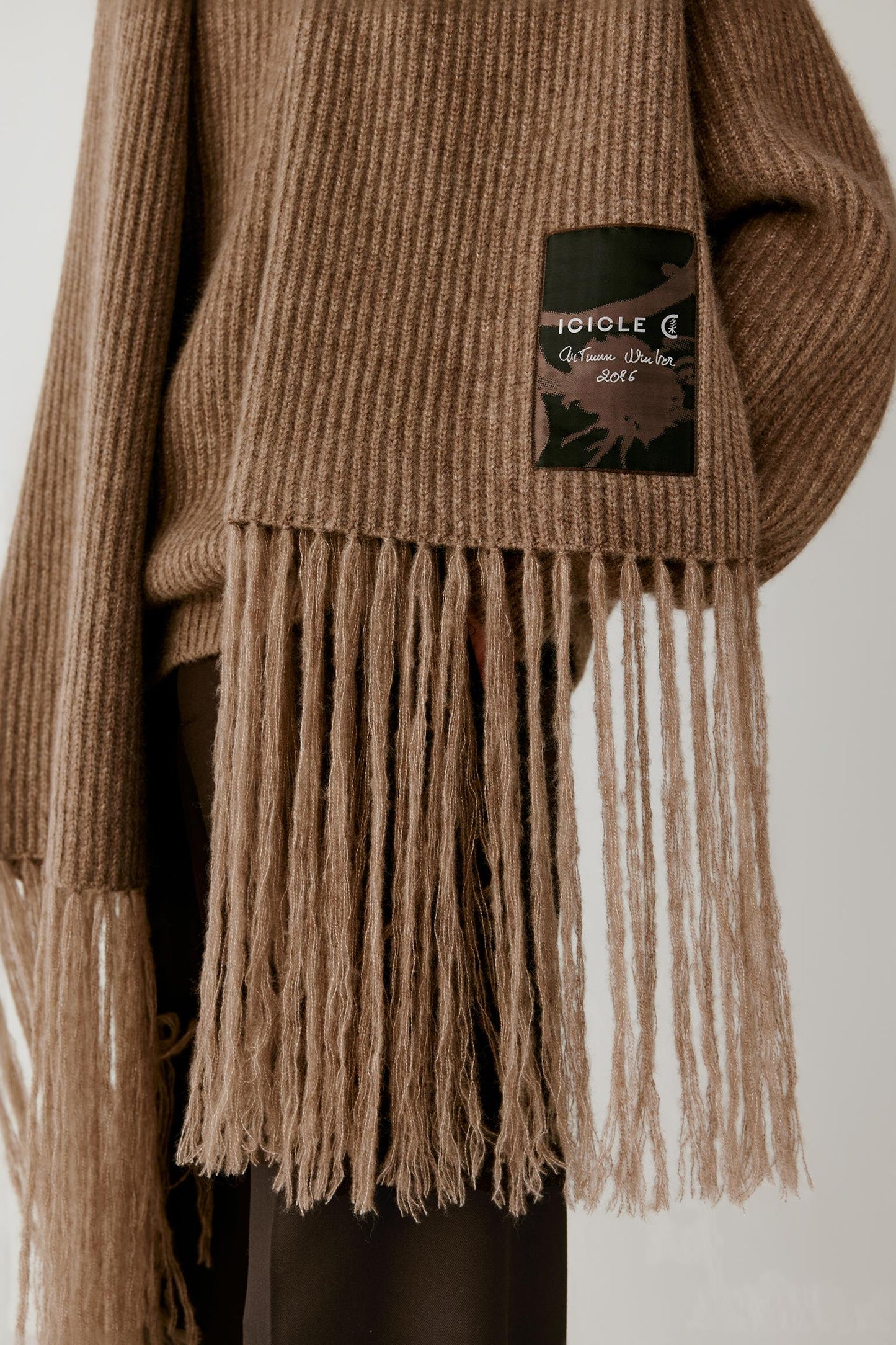 Close-up of a ribbed knit sweater in a warm taupe-brown color, featuring a long fringe hem and a black brand patch on the sleeve with the text 'JOULE C Collection Cotonnet'.