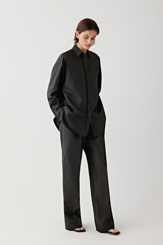 Full-length studio photograph of a woman wearing a black shirt made from gambiered Canton gauze silk. The shirt has a straight relaxed silhouette with long sleeves, a classic collar and a buttoned front closure. It is worn with matching trousers.