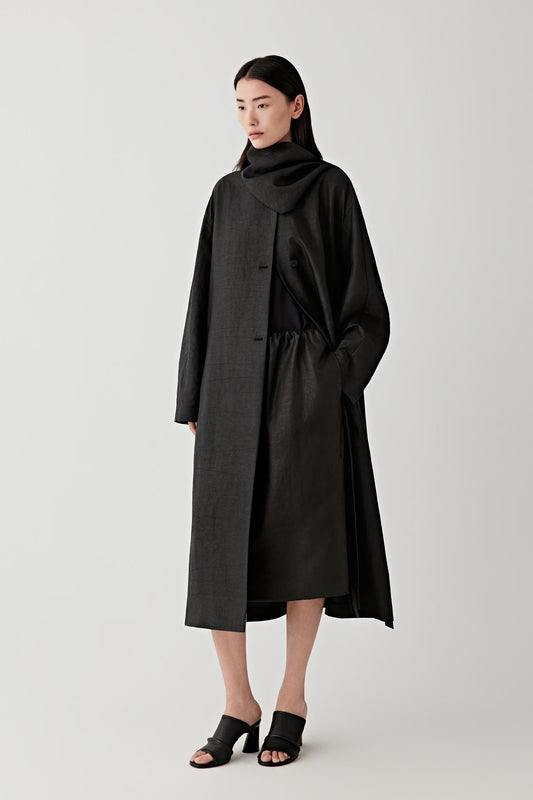Front view of a long black trench coat in gambiered Canton gauze worn by a woman. The coat features a relaxed, oversized cut with wide sleeves, a soft collar, and a belt that gathers the fabric slightly at the waist while maintaining a loose drape.