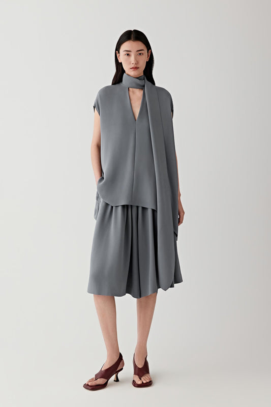 Full-length front view showing the elongated silhouette and wrap-style neckline extending into a front panel construction. The sleeveless cut and fluid silk crepe create soft movement while maintaining clean vertical lines.