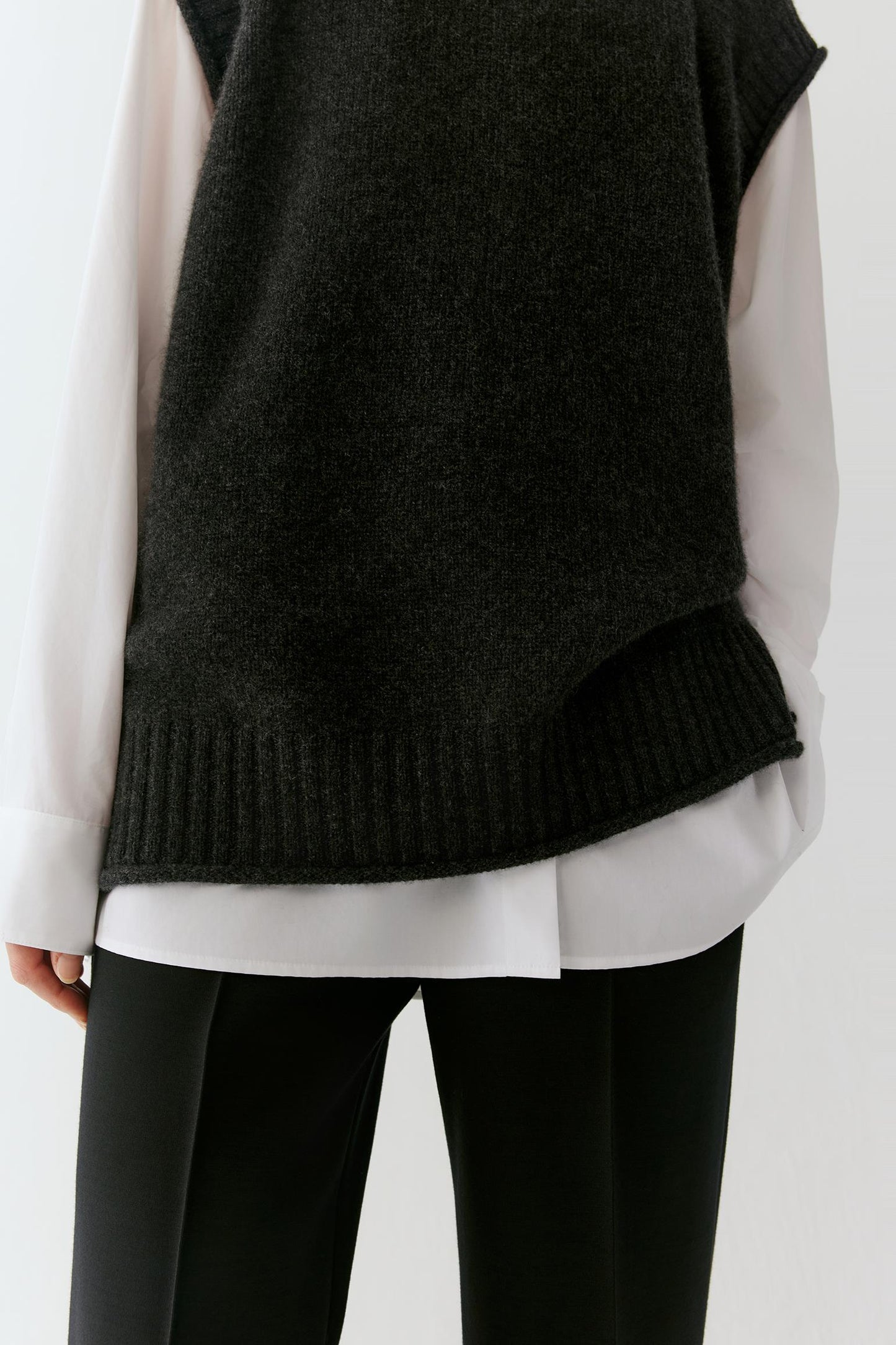 A close-up of a person wearing a dark charcoal knit sleeveless sweater vest over a white long-sleeve shirt and black pants.