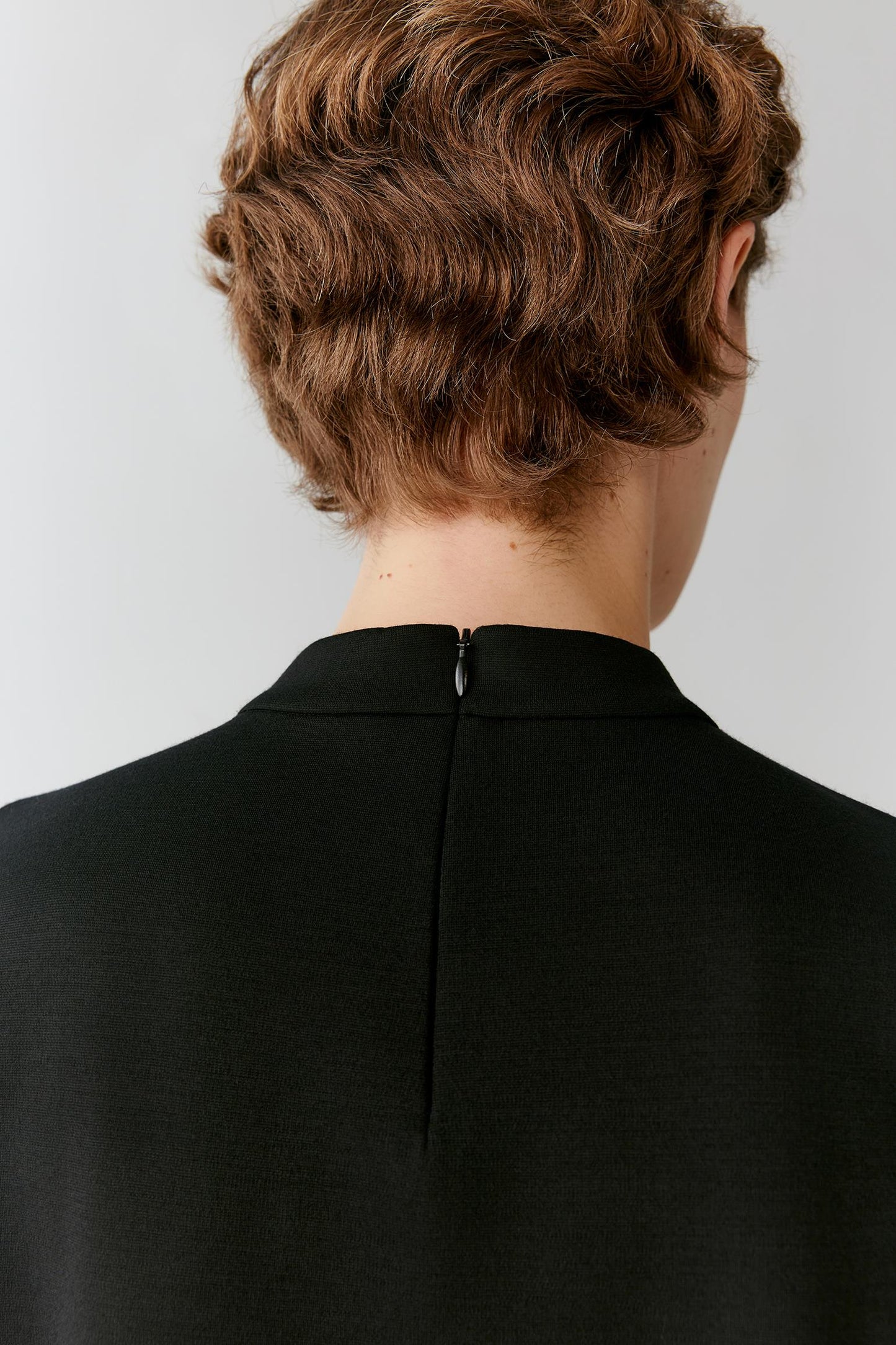 Back view of a person with short, wavy brown hair wearing a black high-neck top or dress, showing the center back zipper against a light gray studio background.