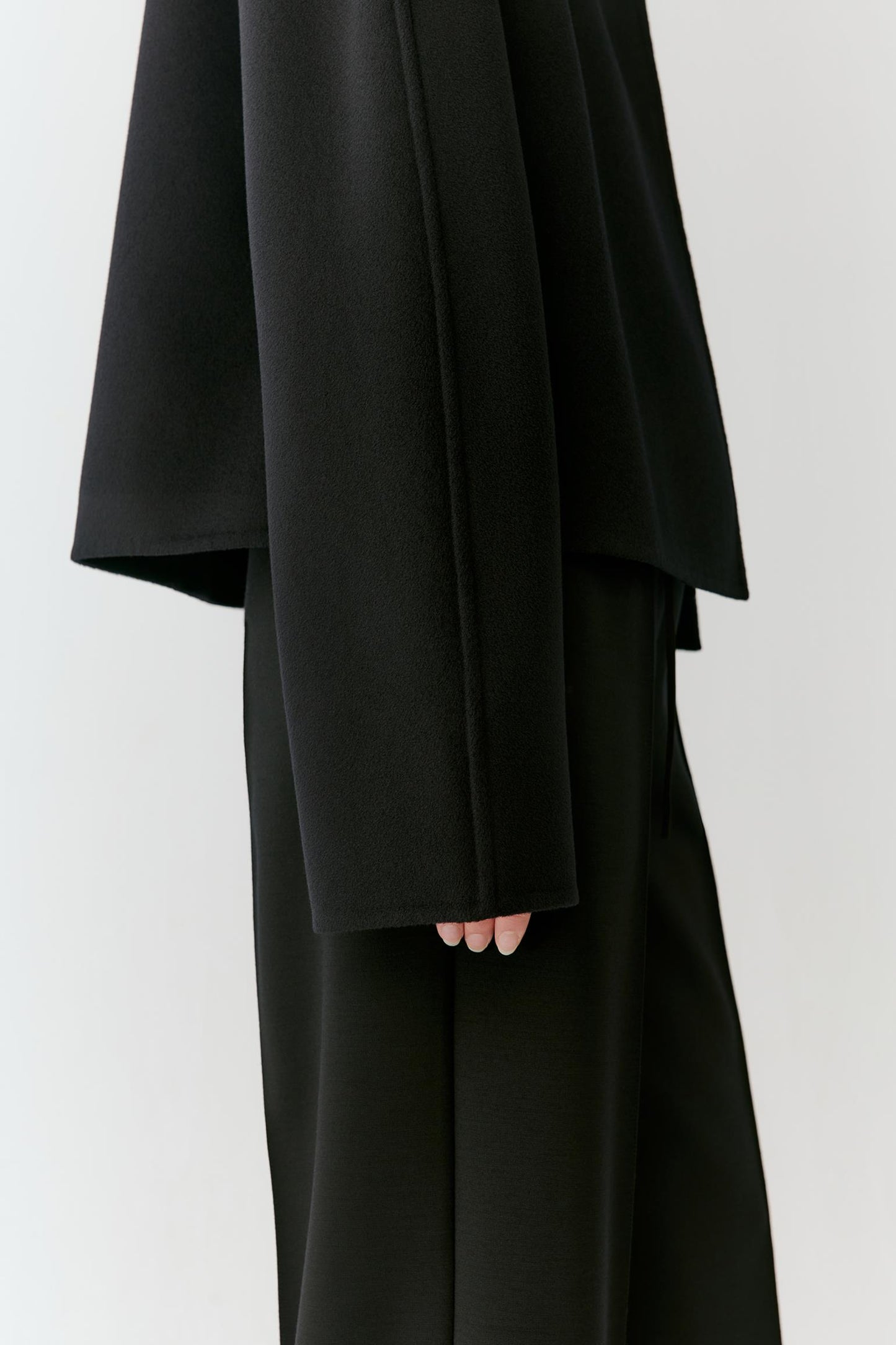 A close-up of a person wearing a black draped, oversized long-sleeve top or dress with matching black wide-leg bottoms, shown against a plain light gray background.