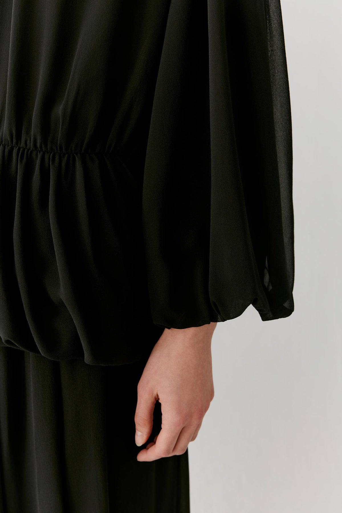 Close-up of a person wearing a black sheer long-sleeve top with a gathered waist and a visible hand, against a plain light gray background.