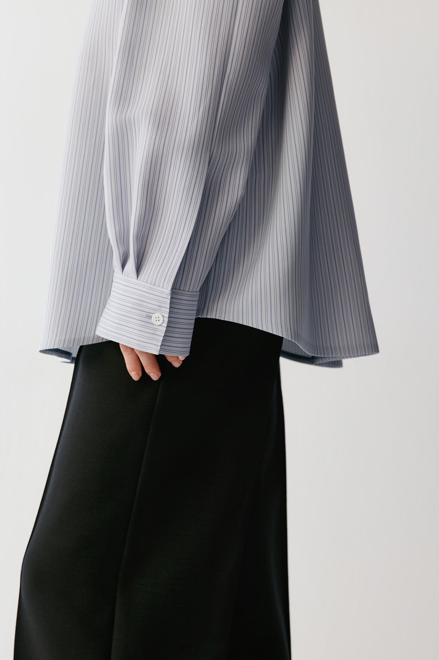 A close-up of a person wearing a light gray and white vertically striped long-sleeve blouse with a buttoned cuff, paired with black wide-leg trousers, against a plain light gray background.