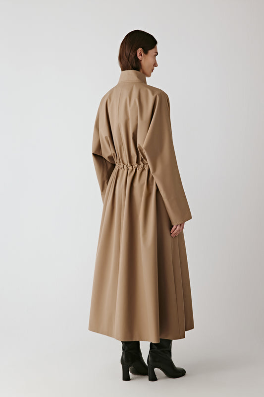 Back view of the long beige coat showing the tied belt and the fluid drape of the fabric.