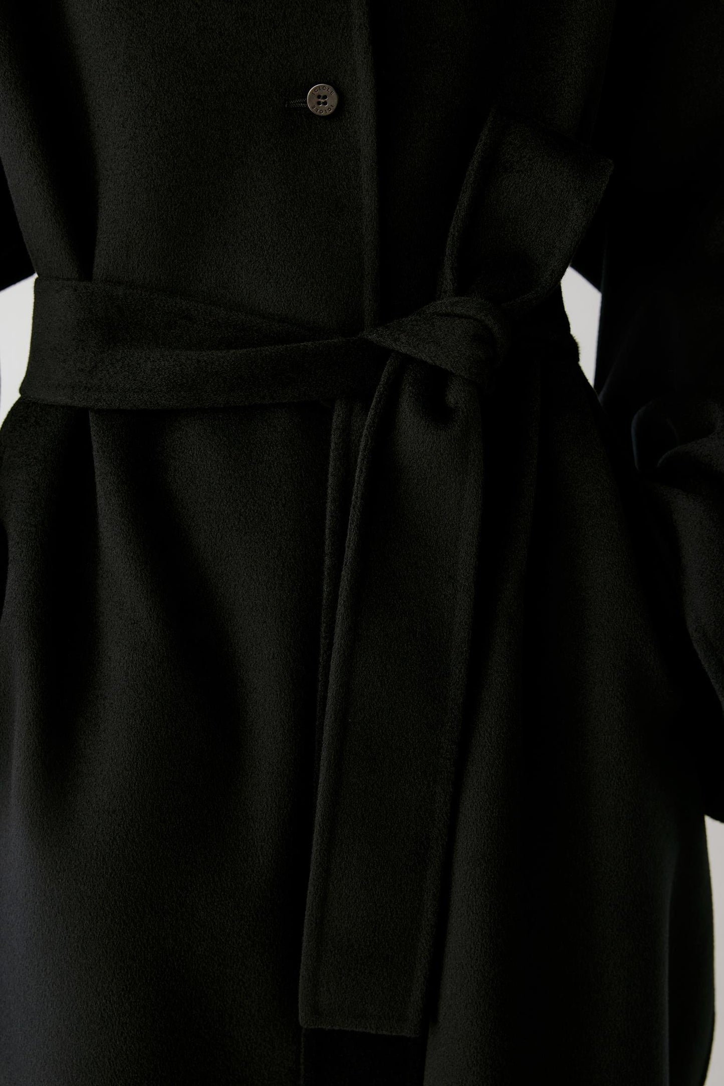 Close-up of a black belted coat with a single visible button and soft fabric folds, photographed against a dark background.