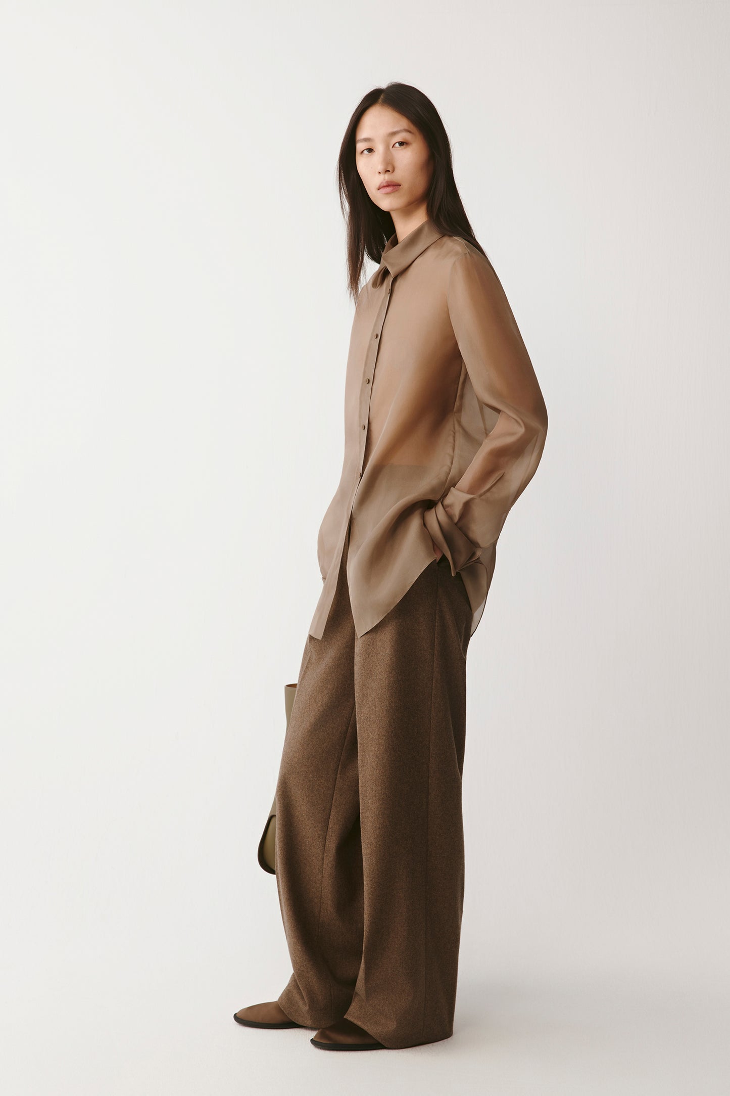 Three-quarter view of a model wearing a light brown silk organza shirt paired with wide brown wool trousers. The sheer fabric subtly reveals the silhouette beneath, highlighting the shirt’s fluid drape and delicate transparency.