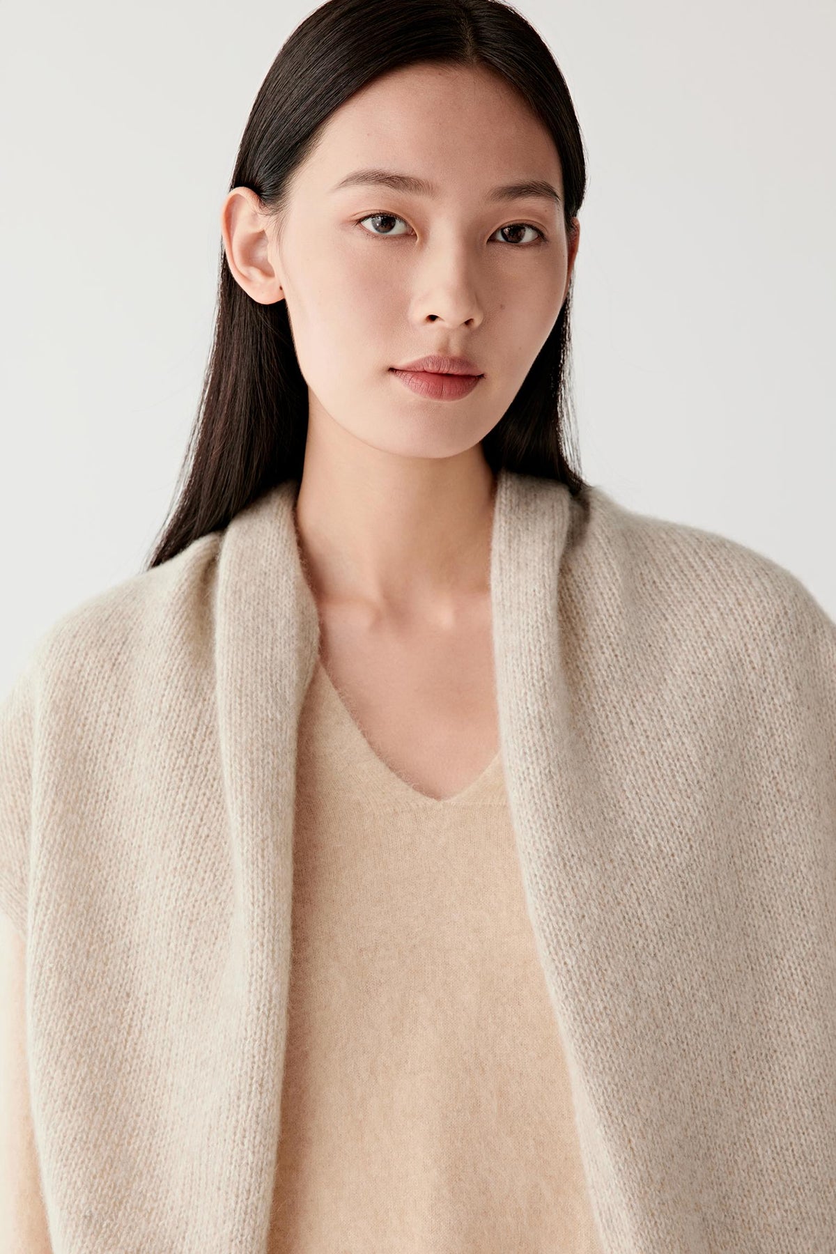 A woman wearing a soft beige knit cardigan draped over a matching beige V-neck sweater, photographed against a clean light gray studio background.