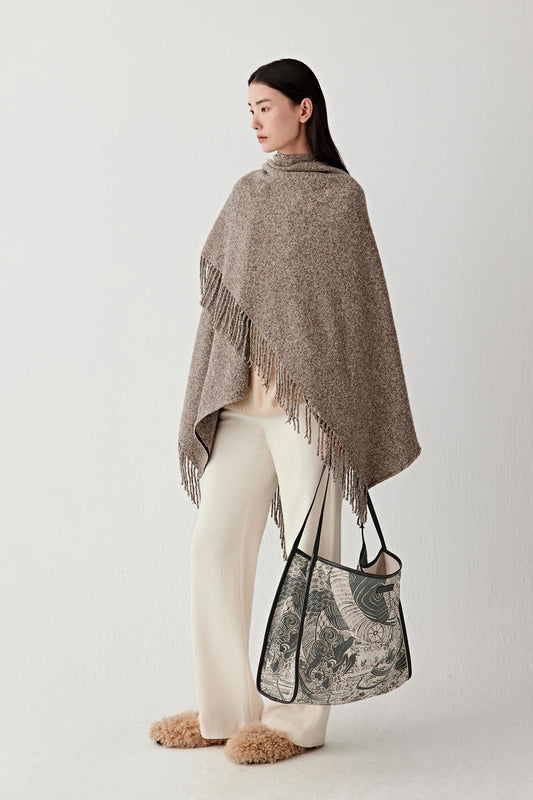 Woman wearing a brown poncho with fringe, holding a patterned bag on a white background