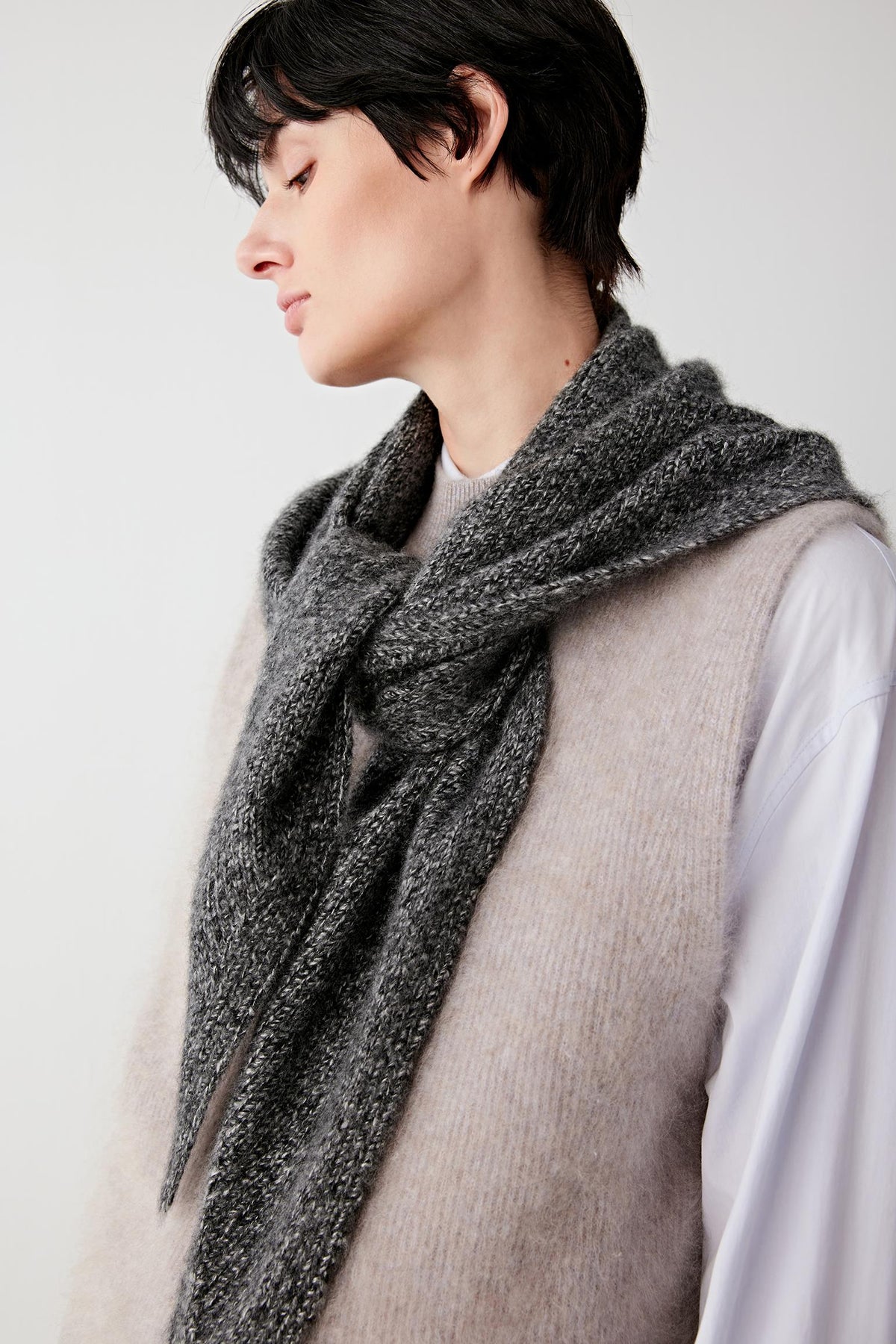 A close-up side profile of a person wearing a thick, textured dark gray knit scarf wrapped around a light beige knit vest over a white shirt, against a plain light gray background.