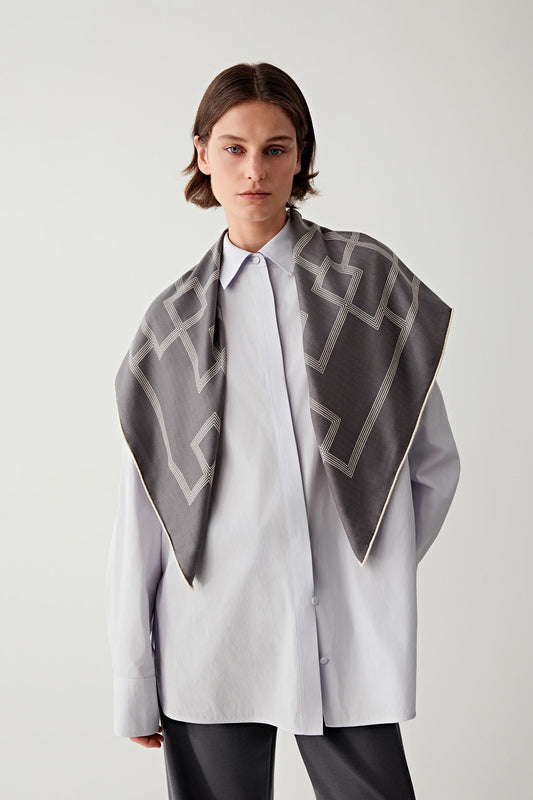 Model wearing a square cashmere and silk scarf in soft grey tones, draped over the shoulders in a triangular shape, showing the geometric pattern and fluid drape against a light shirt.