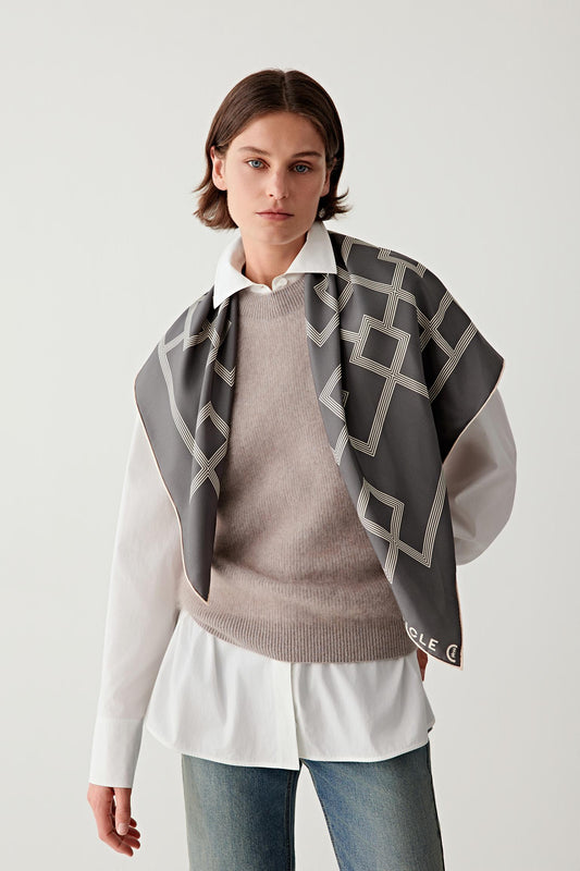 Front view showing the scarf worn loosely around the neck over a light beige sweater. The structured geometric pattern becomes visible as the silk twill folds naturally.