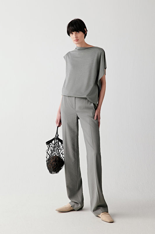 Full-length front view showing the relaxed silhouette shaped by a draped collar, open shoulders and asymmetrical sleeves. The stretch jersey construction follows the movement of the body while maintaining a soft and fluid line.