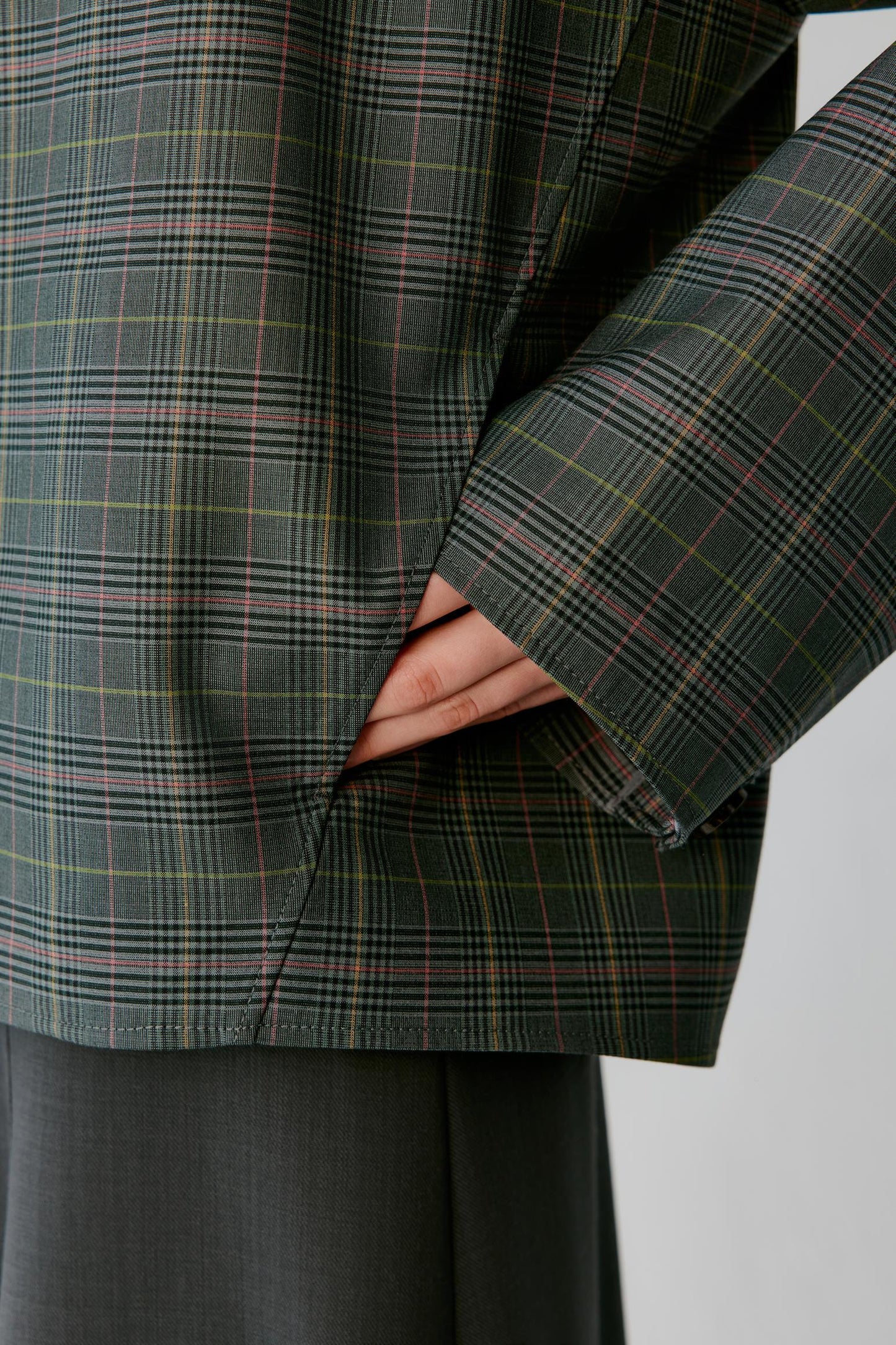 Close-up of a person wearing a dark green plaid blazer with thin red and yellow lines, showing the fabric texture and a hand tucked into the side pocket, paired with dark gray trousers against a light gray background.