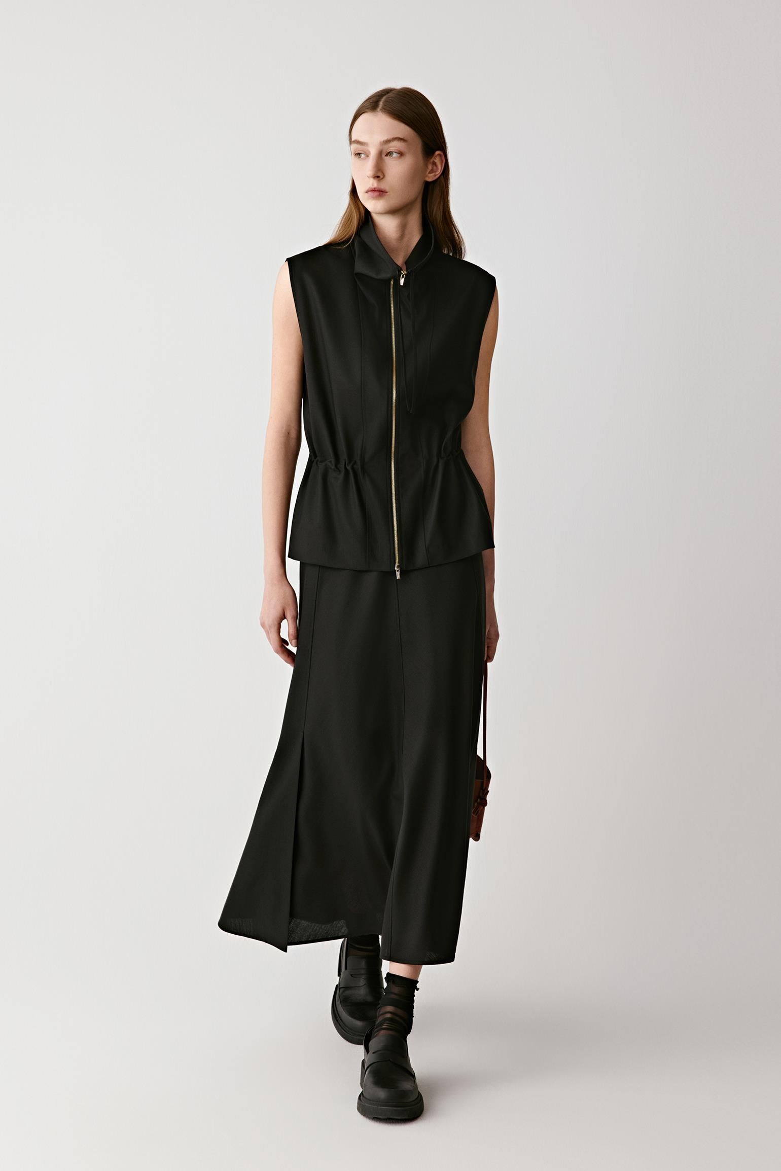 Front full-length view of a woman wearing a black sleeveless combed wool jacket over boots for a structured yet fluid look.