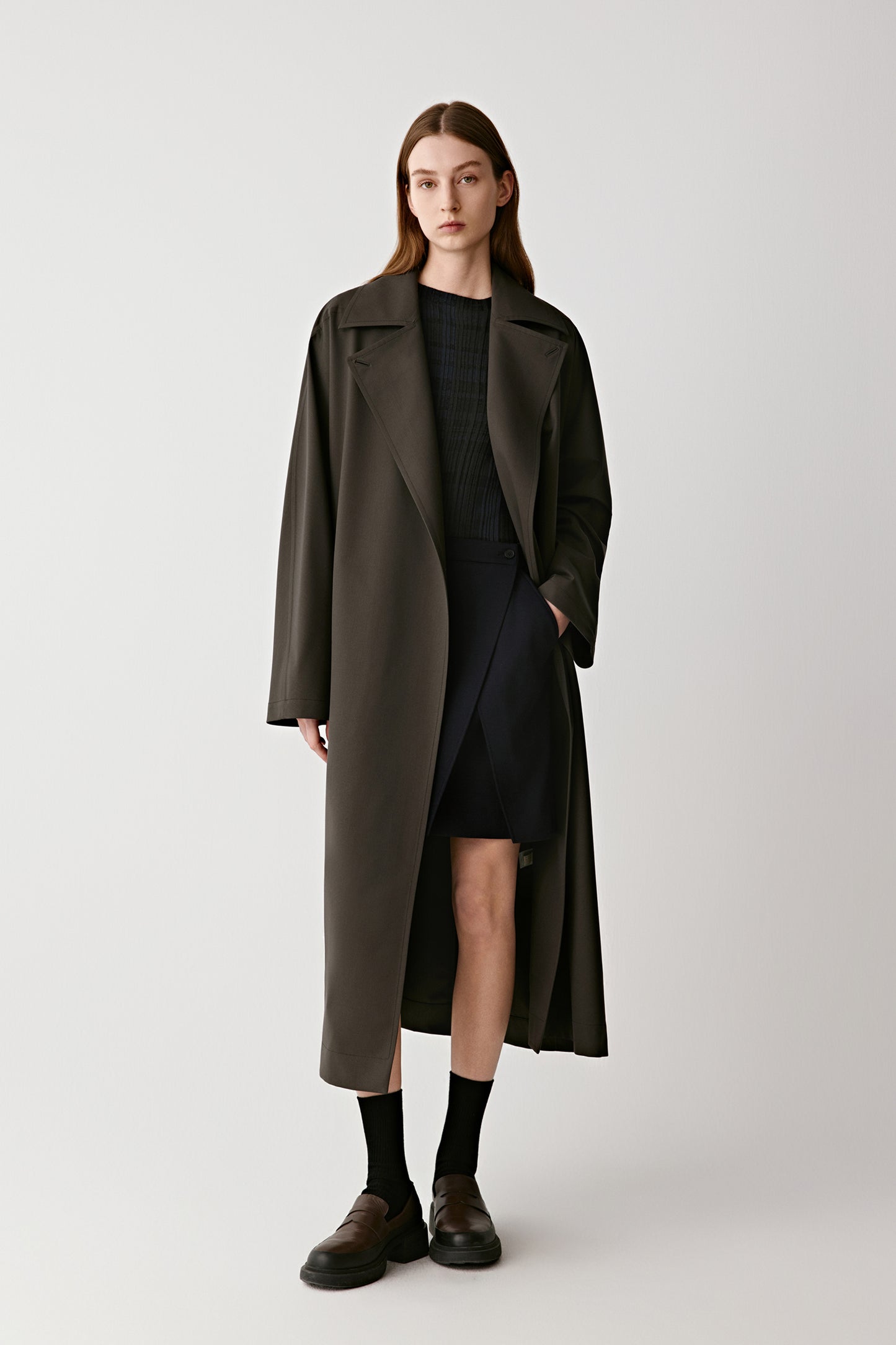 Full-length front view of a woman wearing a dark wool and silk DEW trench coat with a refined straight silhouette.