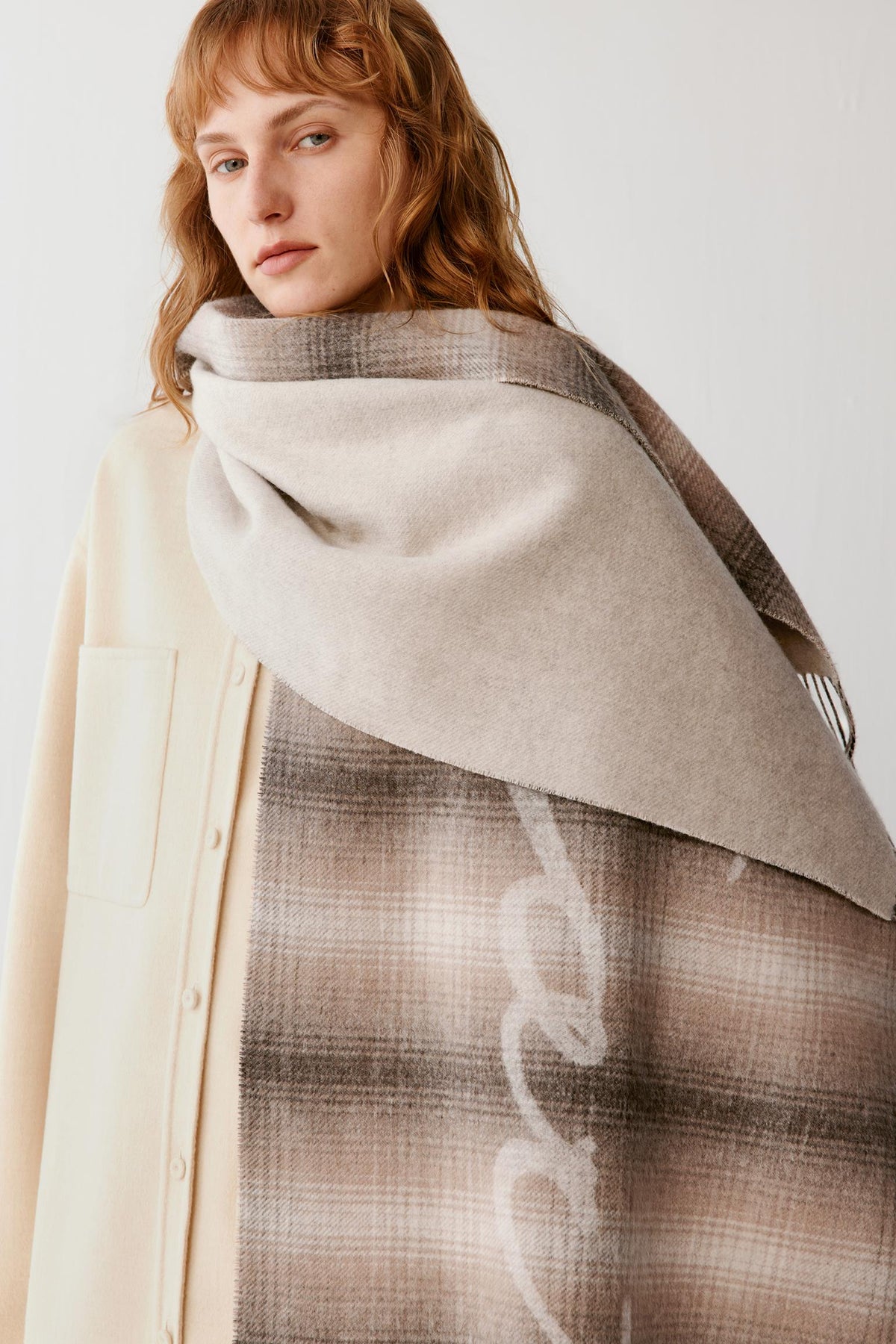 A woman wearing a large beige and brown plaid shawl draped over her shoulders, paired with a cream-colored coat, against a plain light gray background.